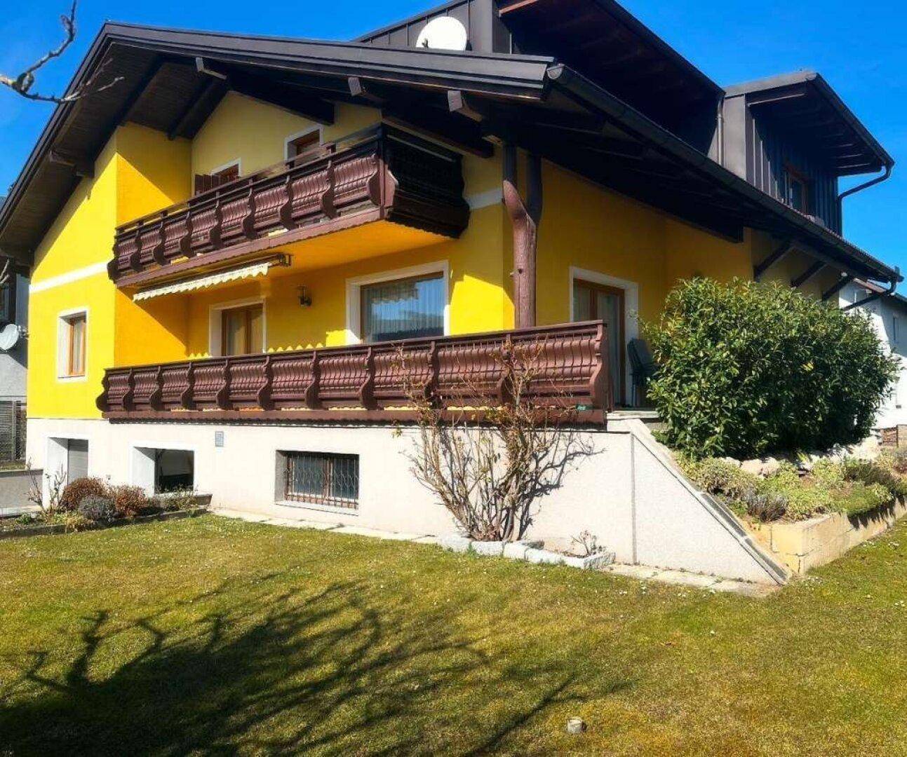 Gelbes Einfamilienhaus mit braunen Balkonen, gepflegtem Vorgarten und Satteldach.