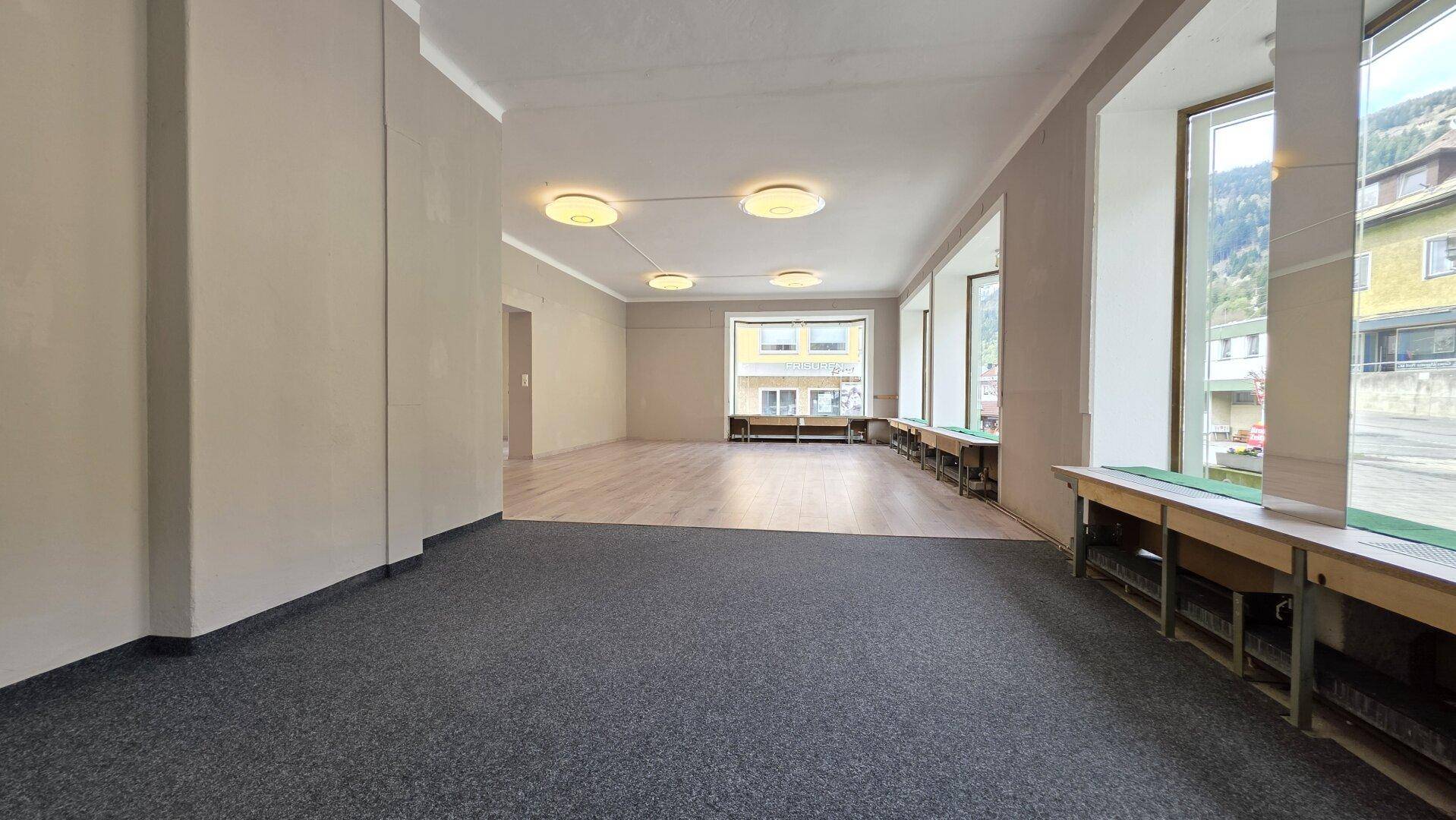 Großer, leerer Raum mit Teppich- und Holzboden sowie breiten Fenstern zur Straße.