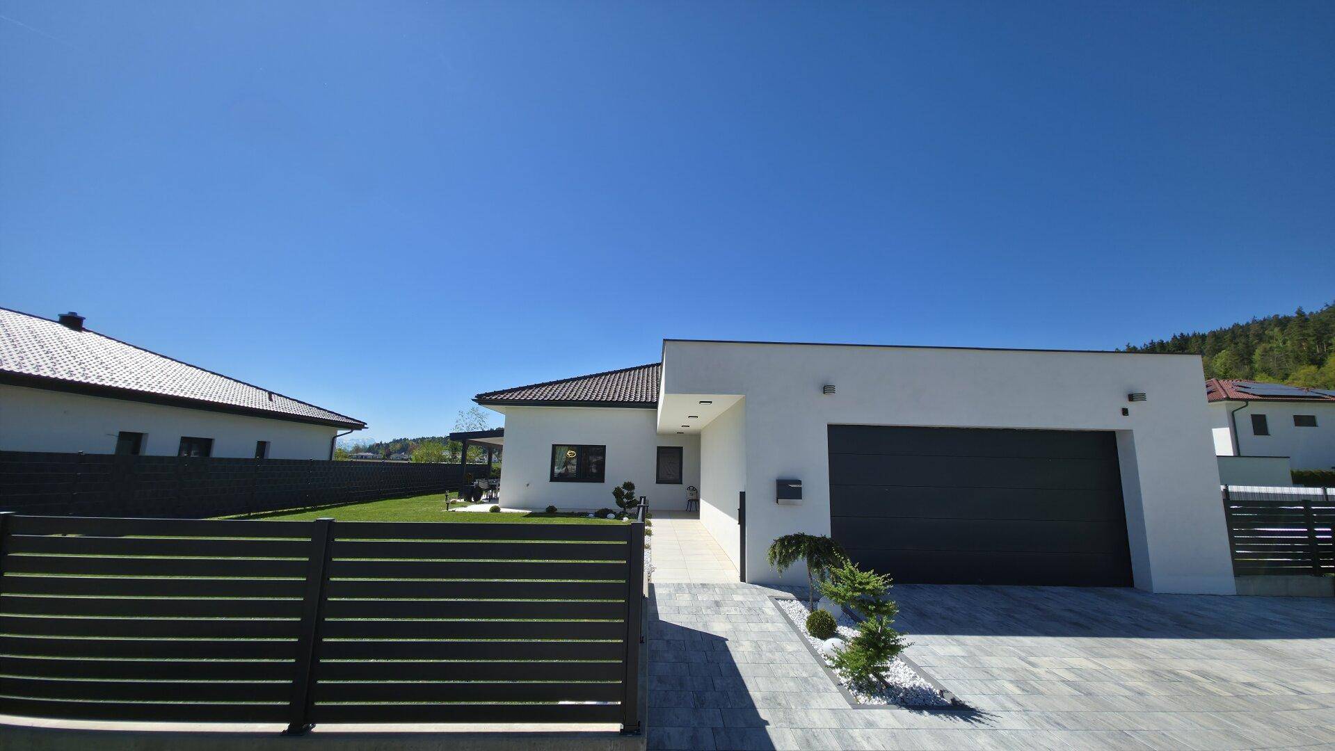 Ansicht der modernen Hausfront mit Garage, Zufahrt und einem eleganten Zaun, umgeben von Grünflächen.