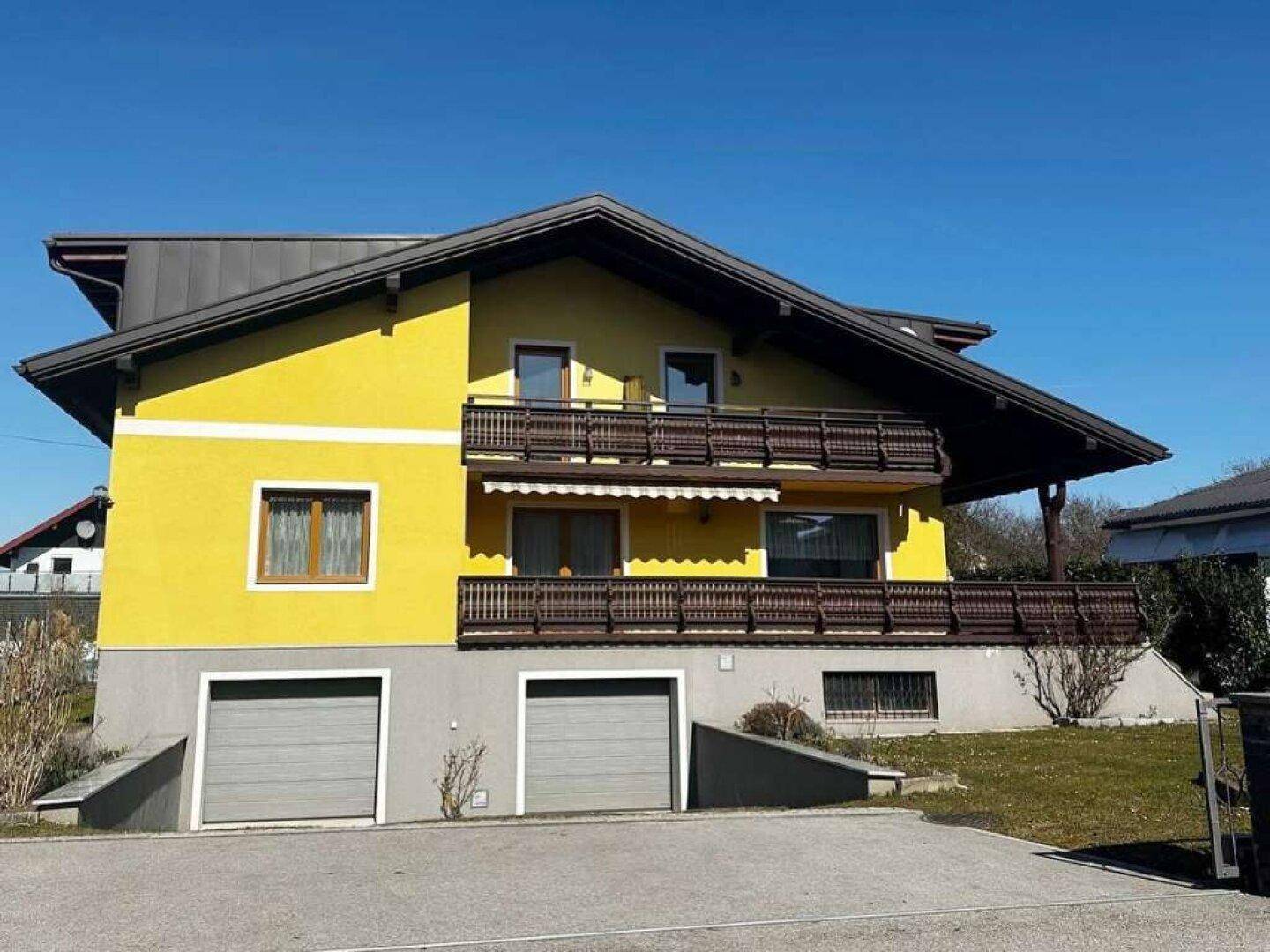 Gelbes Wohnhaus mit zwei Garagen und traditionellen Holzbalkonen unter blauem Himmel.