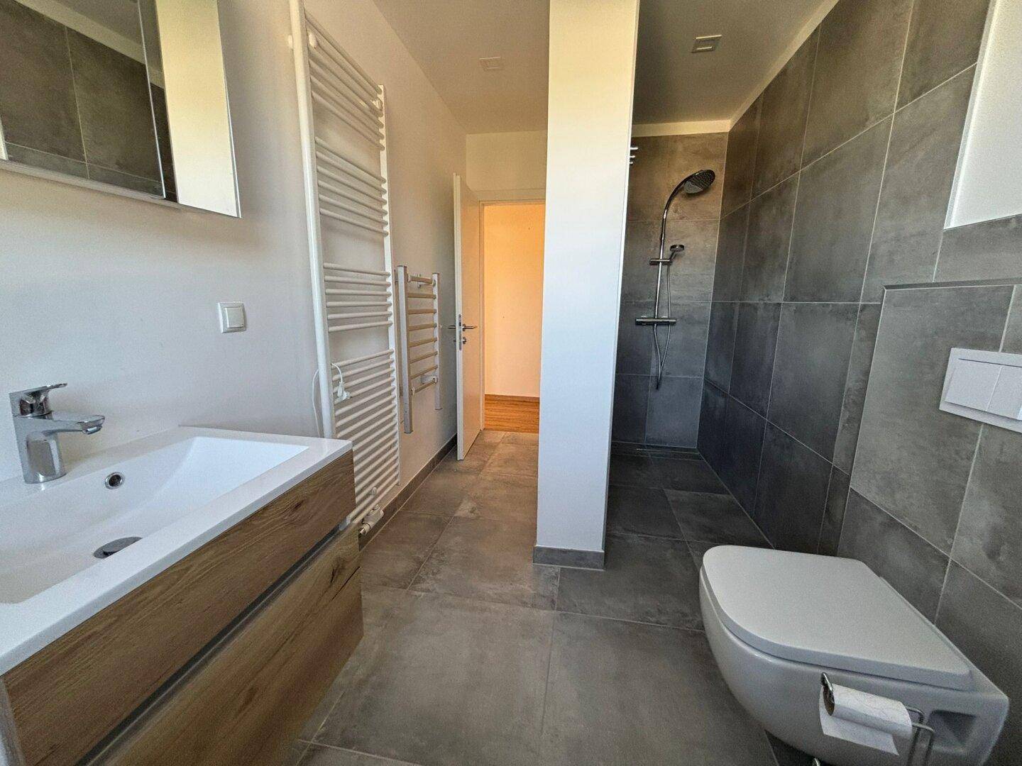 Modernes Badezimmer mit grauen Fliesen, begehbarer Dusche und beheiztem Handtuchhalter.