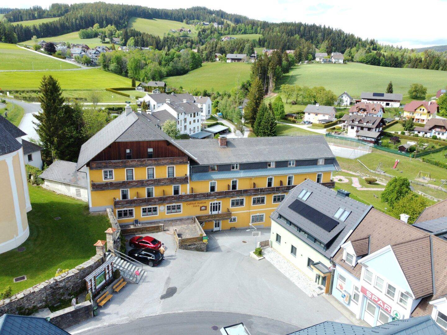Luftaufnahme eines großen Gebäudekomplexes mit Parkplatz und umliegender grüner Landschaft.