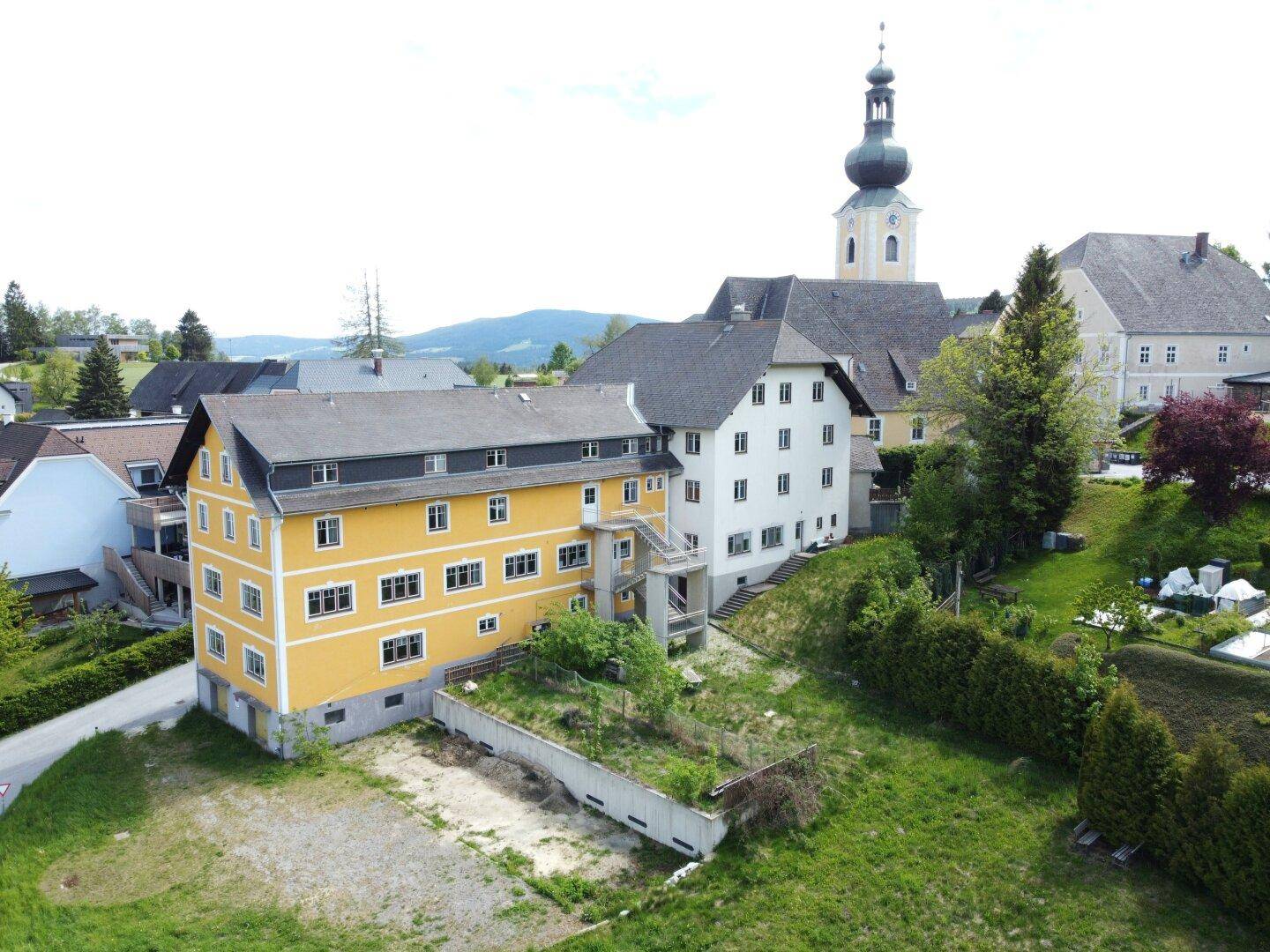 Luftaufnahme eines gelben Gebäudes mit angrenzendem weißen Gebäude und Kirchturm im Hintergrund.
