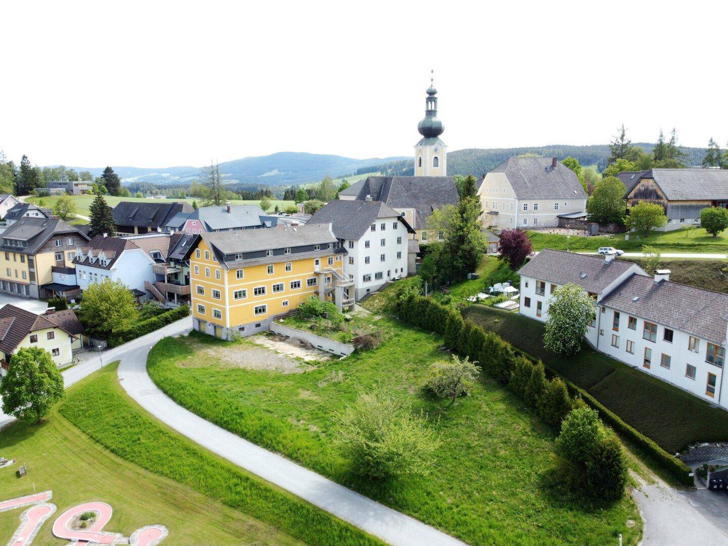 Weitläufige Luftaufnahme eines Gebäudekomplexes in einer grünen, ländlichen Umgebung mit Kirchturm.