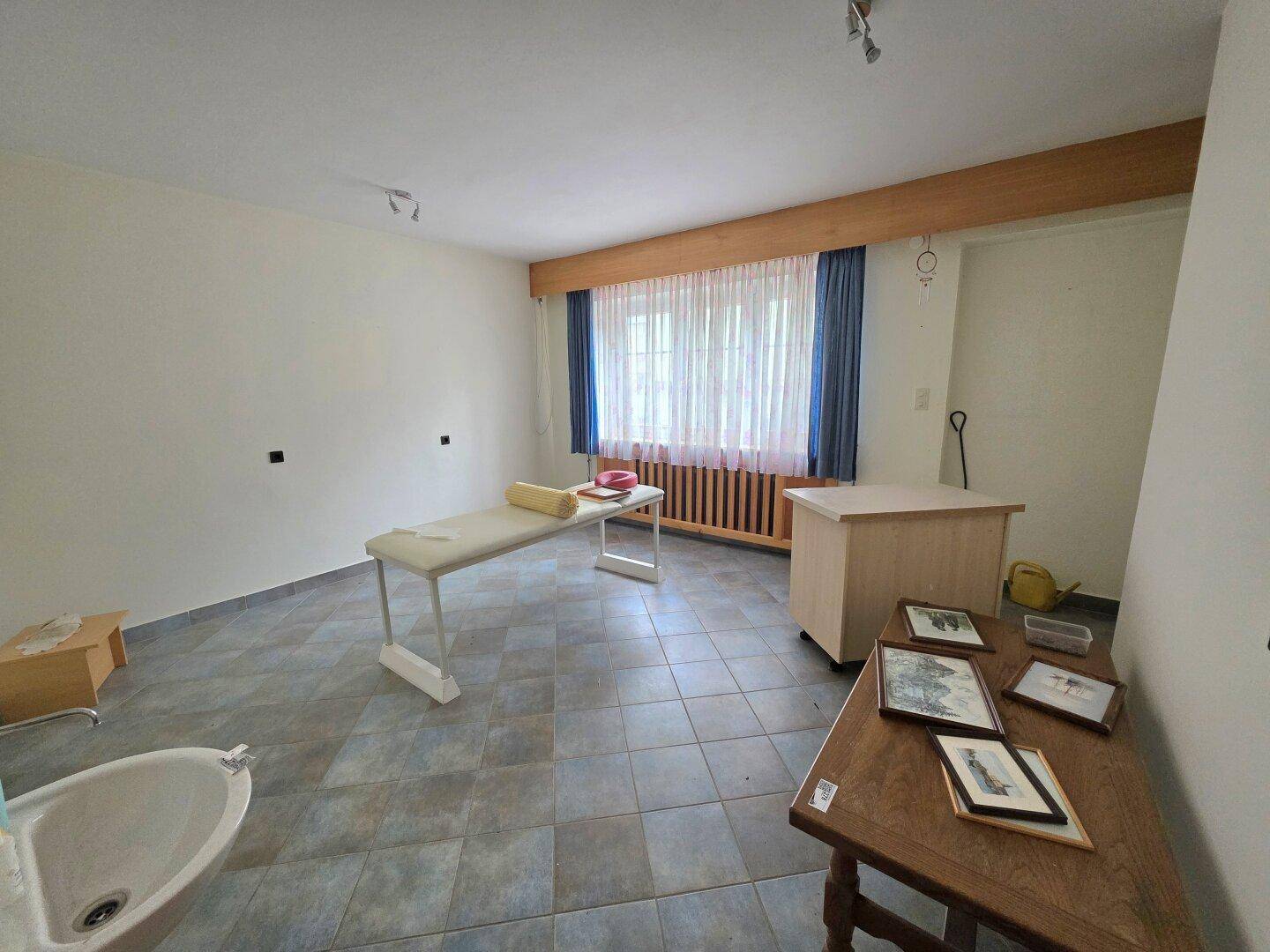 Einfacher Raum mit Fliesenboden, einem Fenster und einer Liege, möglicherweise für Behandlungen.