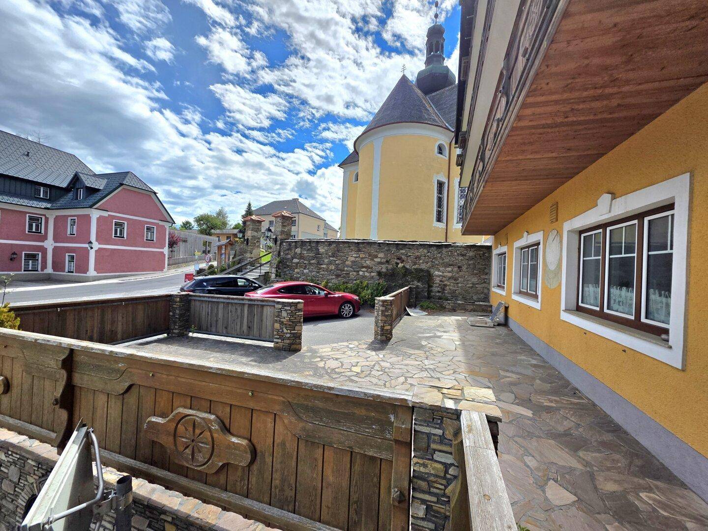 Eingangsbereich eines Gebäudes mit gepflastertem Vorplatz, Holzelementen und Blick auf die Kirche.
