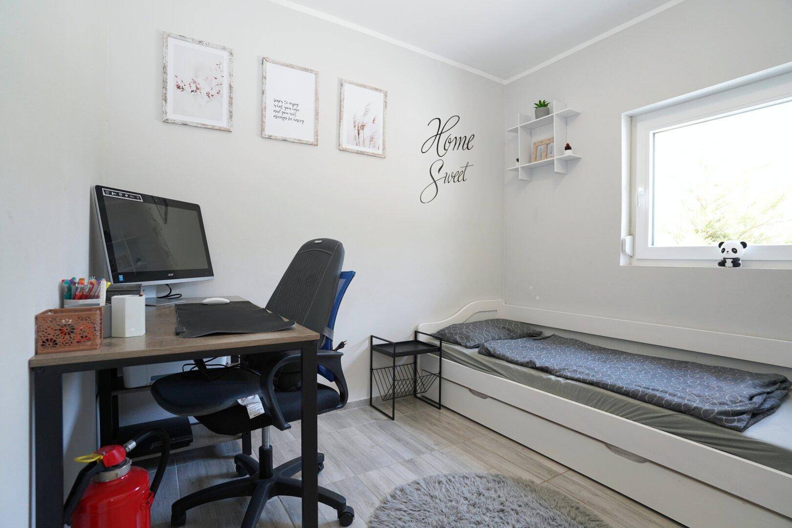 Helles Arbeitszimmer mit Schreibtisch, Einzelbett und Fenster.