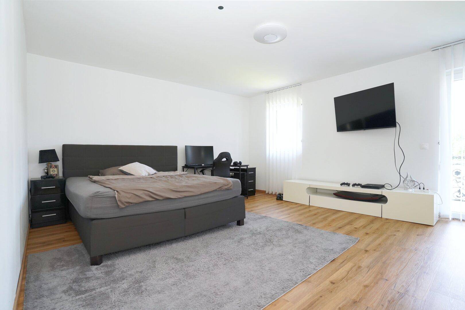 Helles Schlafzimmer mit Doppelbett, Fernseher und Holzoptik-Boden.