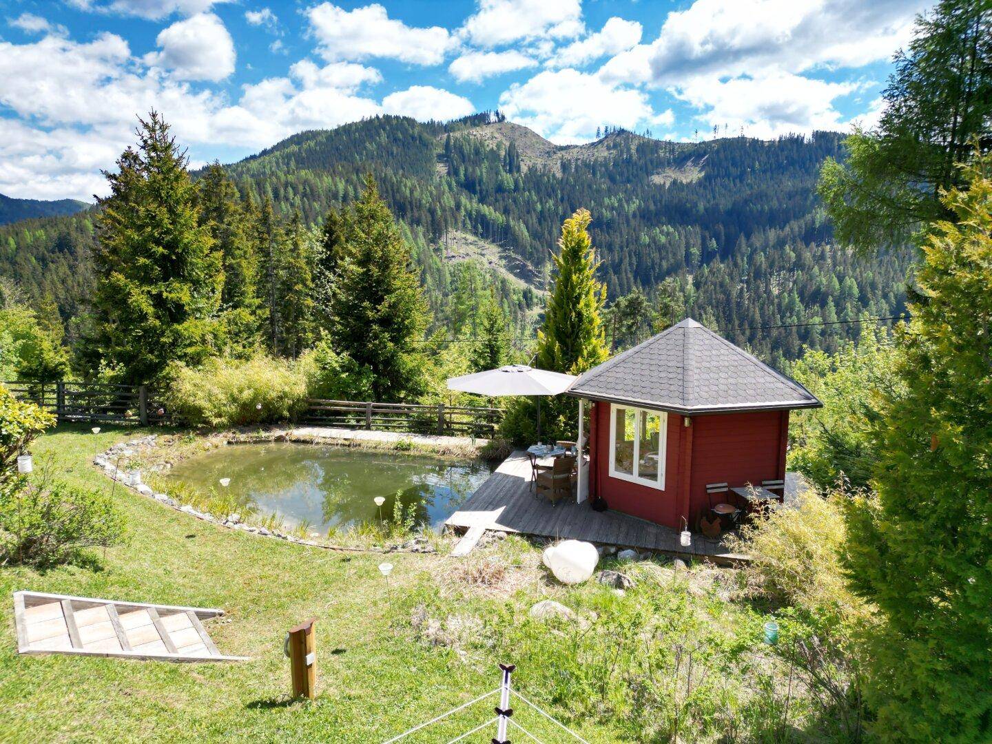 Idyllischer Gartenbereich mit Teich, Holzdeck und einem kleinen Gartenhaus vor Bergkulisse.