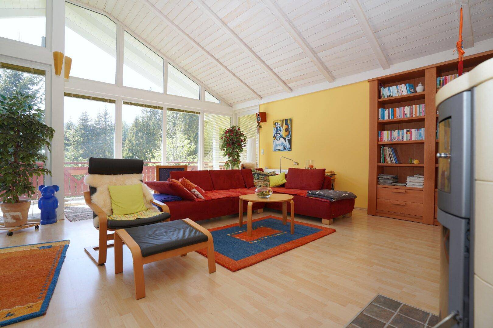 Gemütliches Wohnzimmer mit rotem Sofa, Holzboden und Blick auf die Natur durch große Fenster.