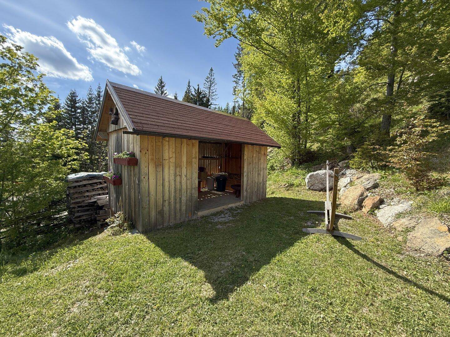 Kleiner Holzschuppen im Garten, umgeben von Grünflächen und Bäumen.