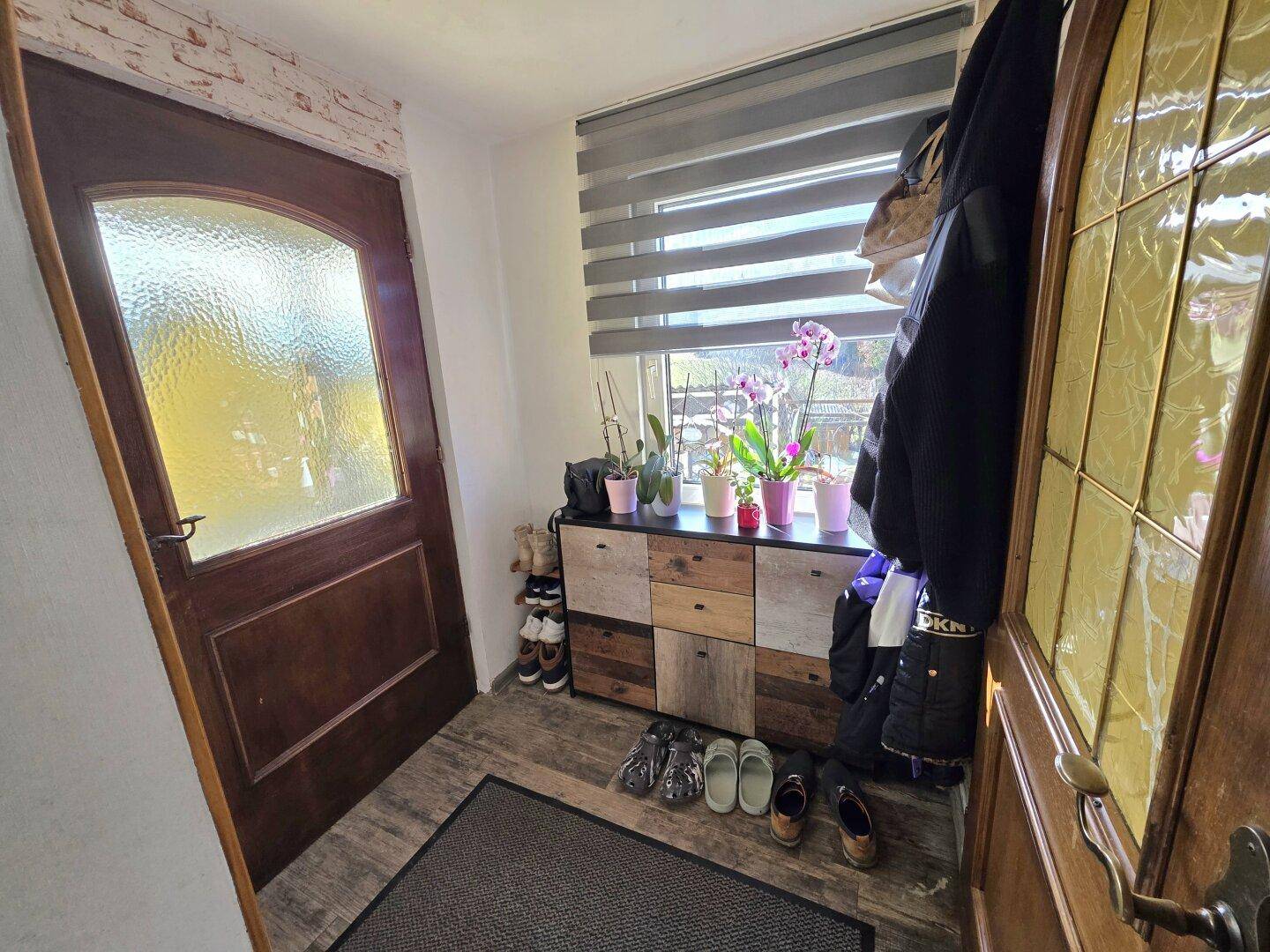 Eingangsbereich mit Holztür, Schuhablage und einem Fenster mit Jalousie.