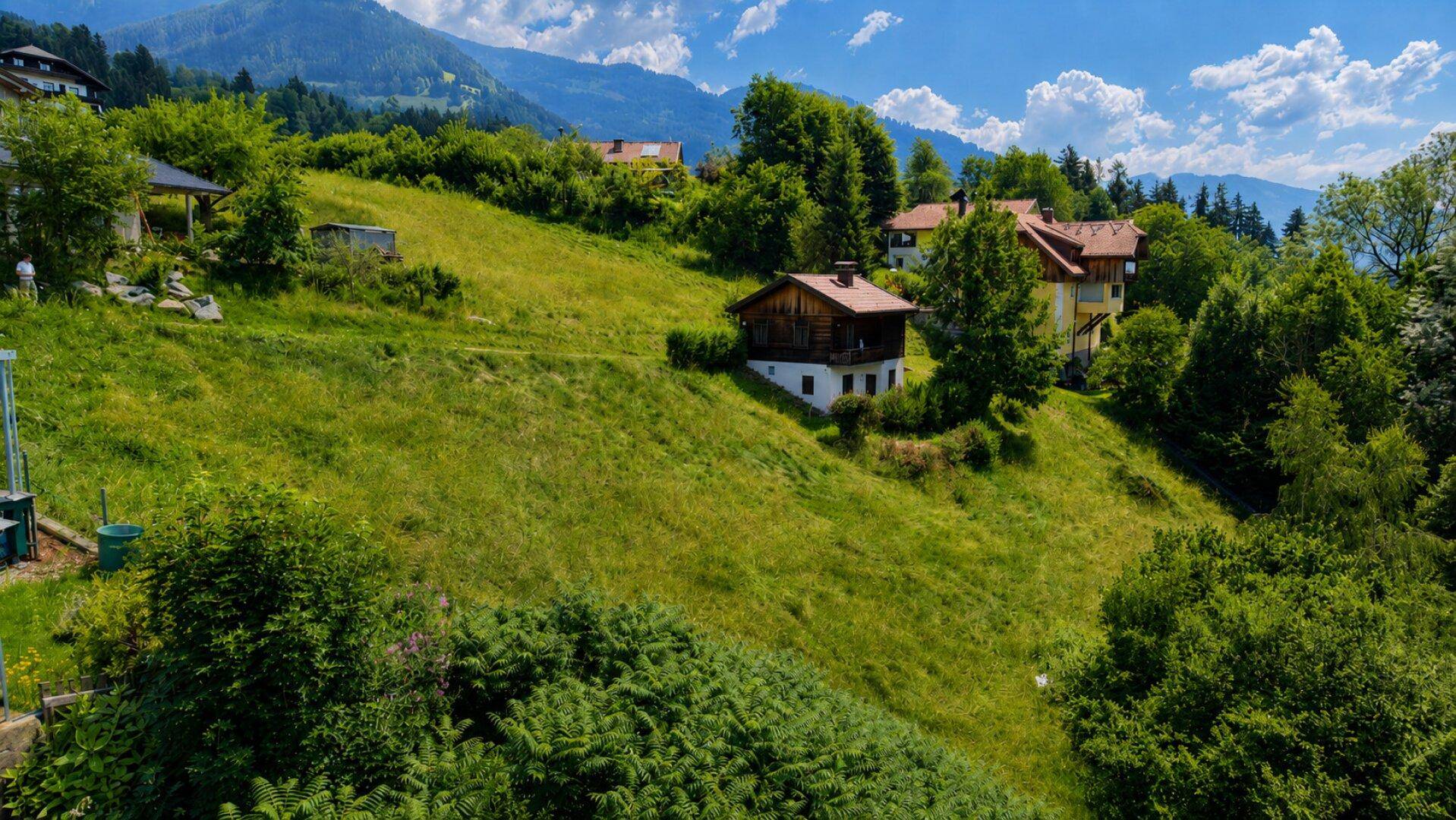 Häuser eingebettet in eine grüne Hügellandschaft mit bewaldeten Bergen im Hintergrund.