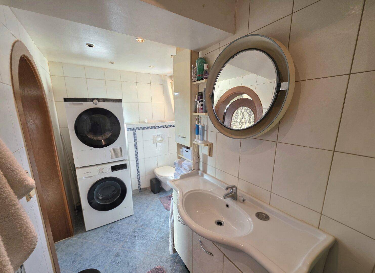 Badezimmer mit Waschmaschine und Trockner, Waschbecken und WC in hellen Fliesen.