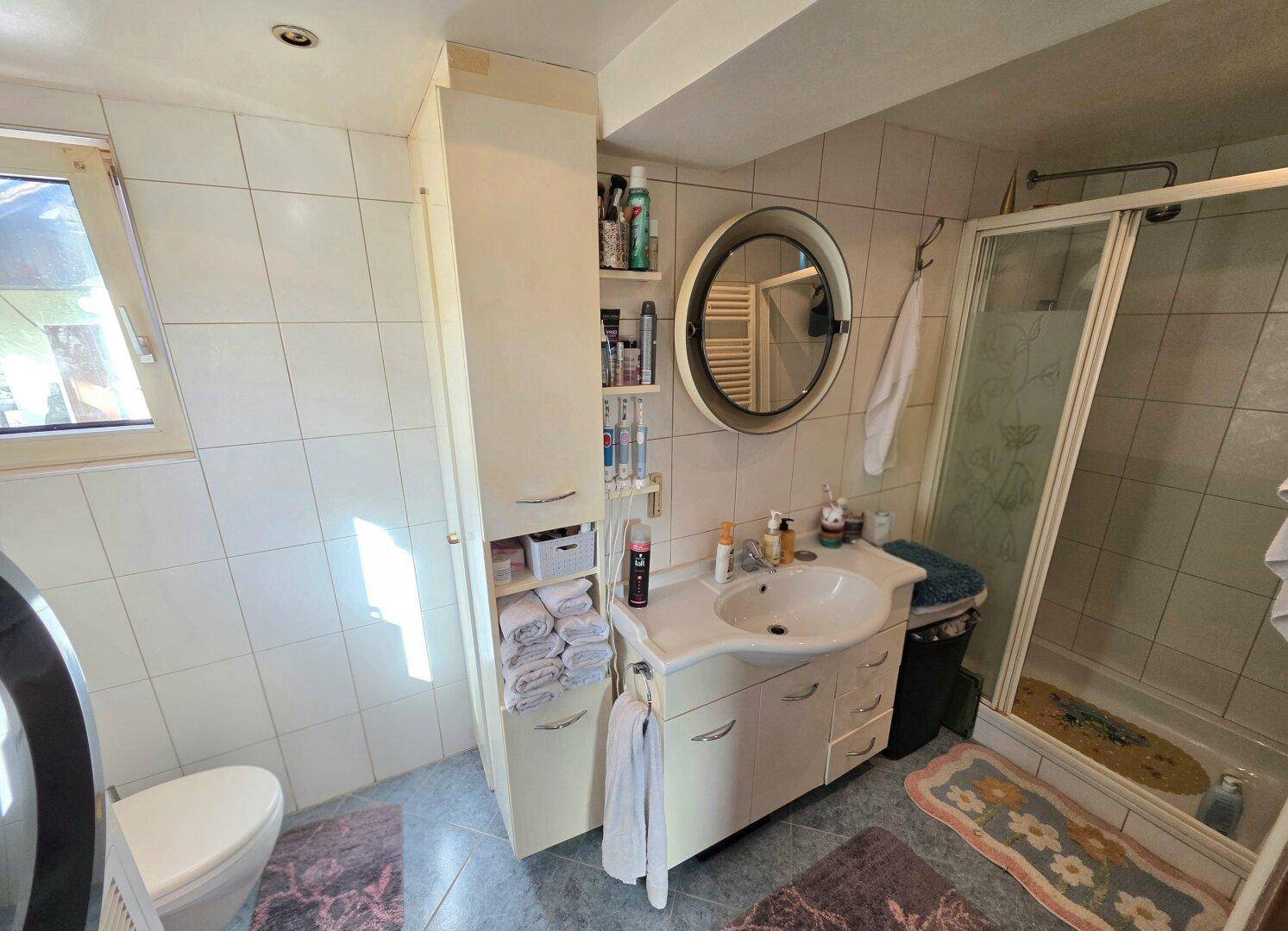 Helles Badezimmer mit Duschkabine, Waschbecken und Fenster in neutralen Fliesen.