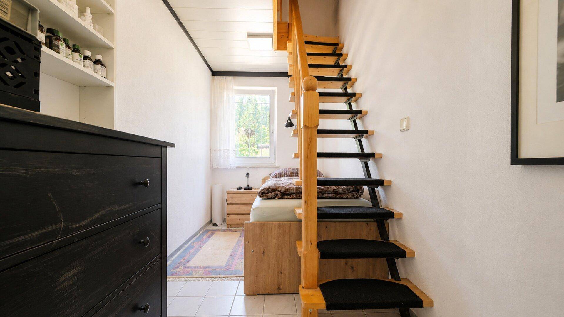 Kleines Schlafzimmer mit Bett, Kommode und einer steilen Holztreppe zum oberen Bereich.