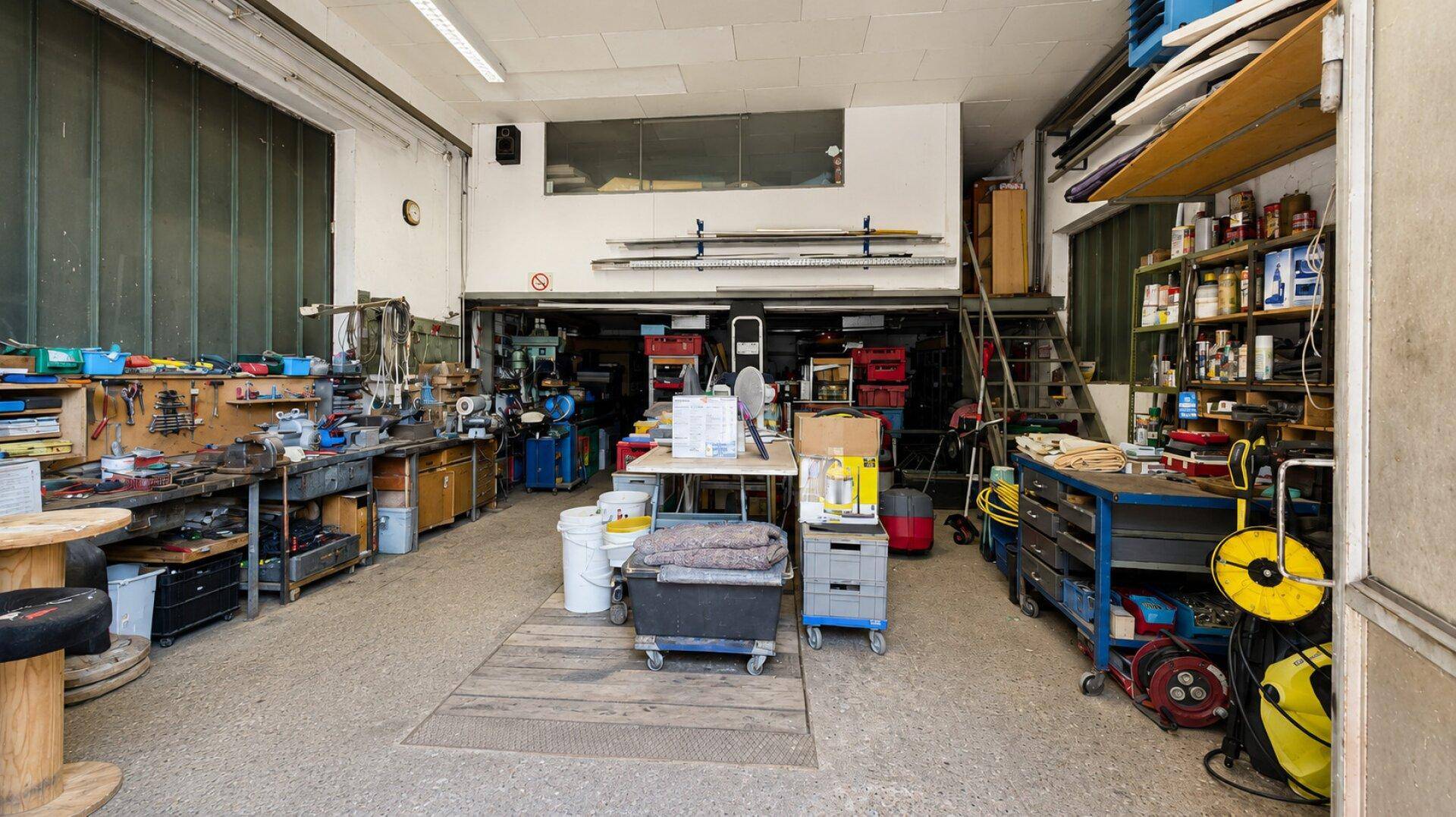 Geräumige Werkstatt mit zahlreichen Werkbänken, Regalen und Arbeitsmaterialien.