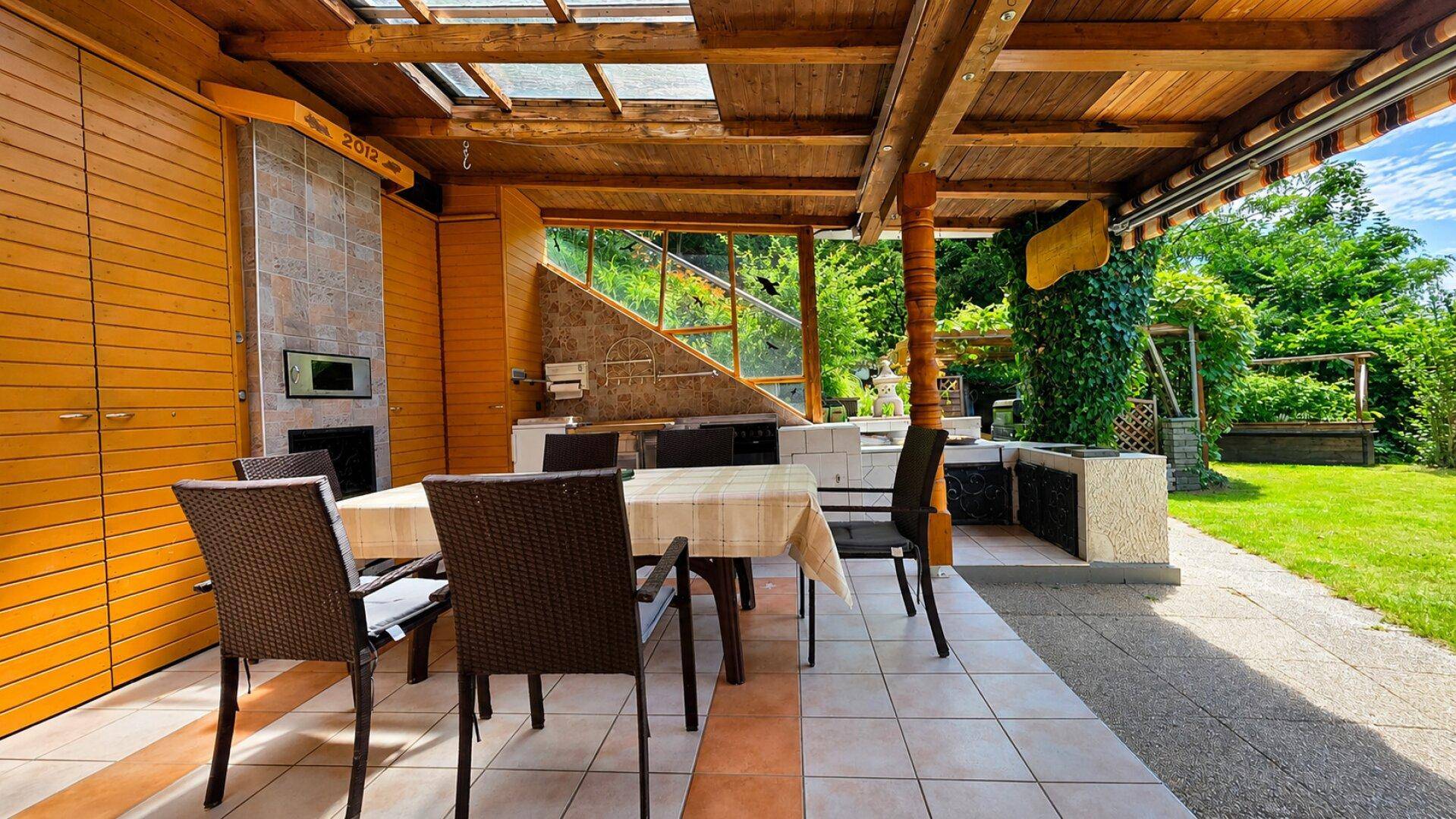 Überdachte Terrasse mit Holzbalken, Fliesenboden und Essbereich im Grünen.
