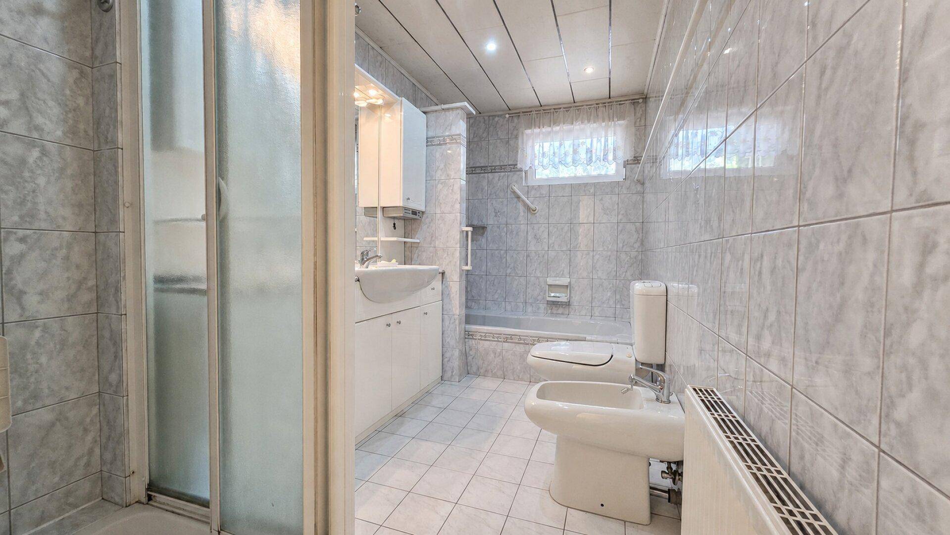 Gefliestes Badezimmer mit Duschkabine, Waschbecken, Toilette, Bidet und Fenster.
