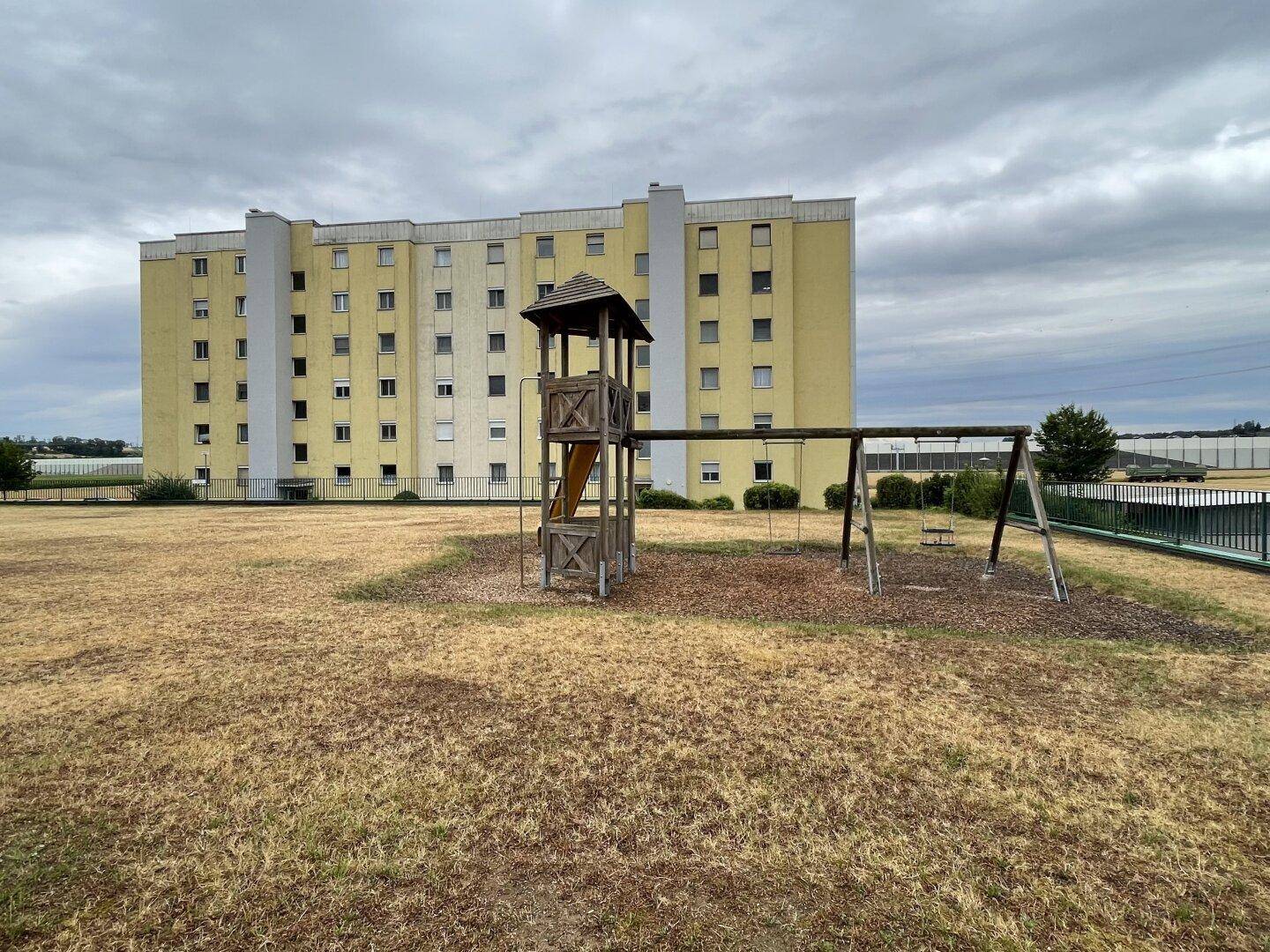 Spielplatz mit Schaukel und Rutsche auf einer Rasenfläche vor einem großen Wohngebäude.