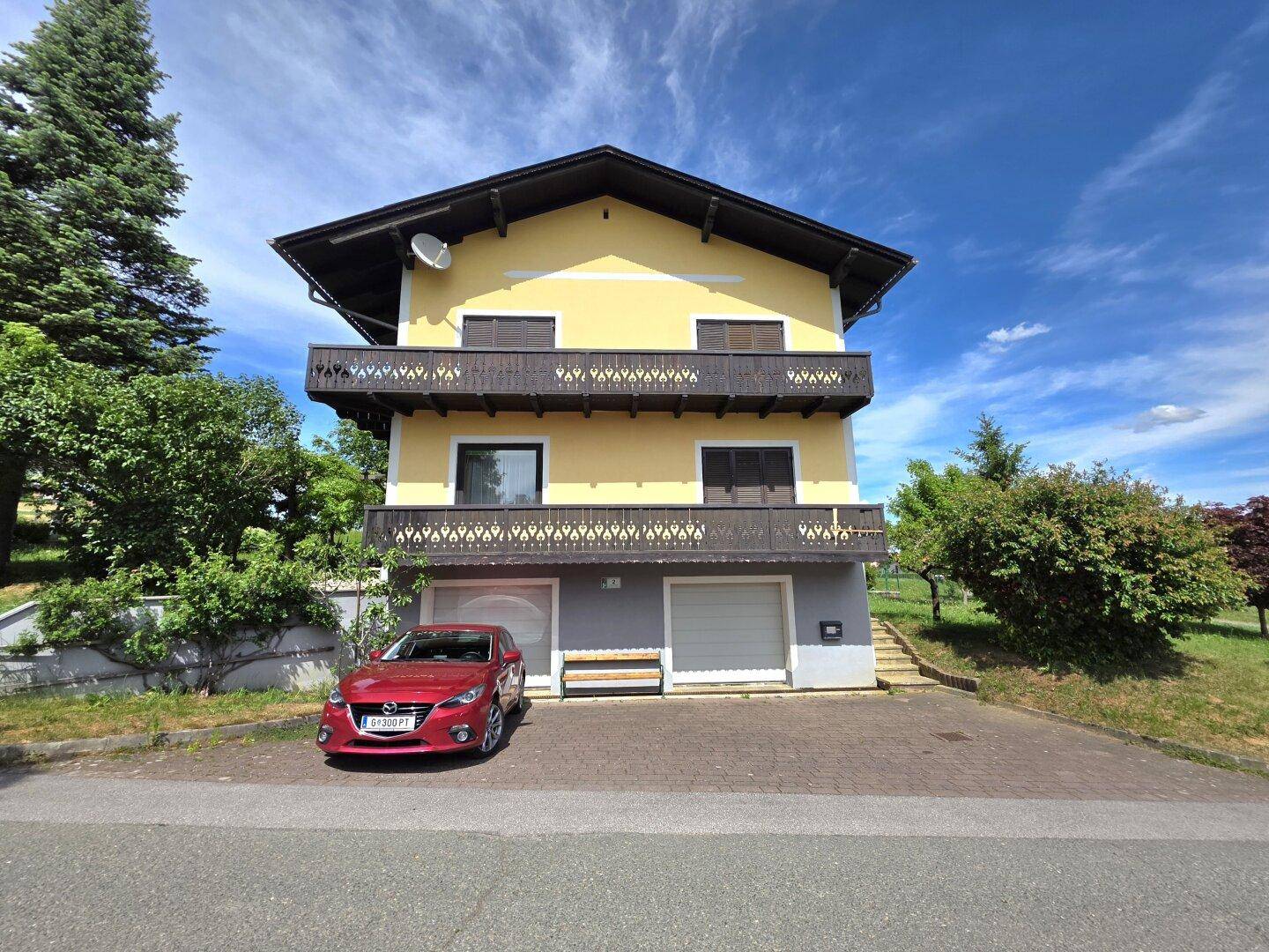 Traditionelles gelbes Haus mit Holzbalkonen, Garagen und gepflasterter Einfahrt.