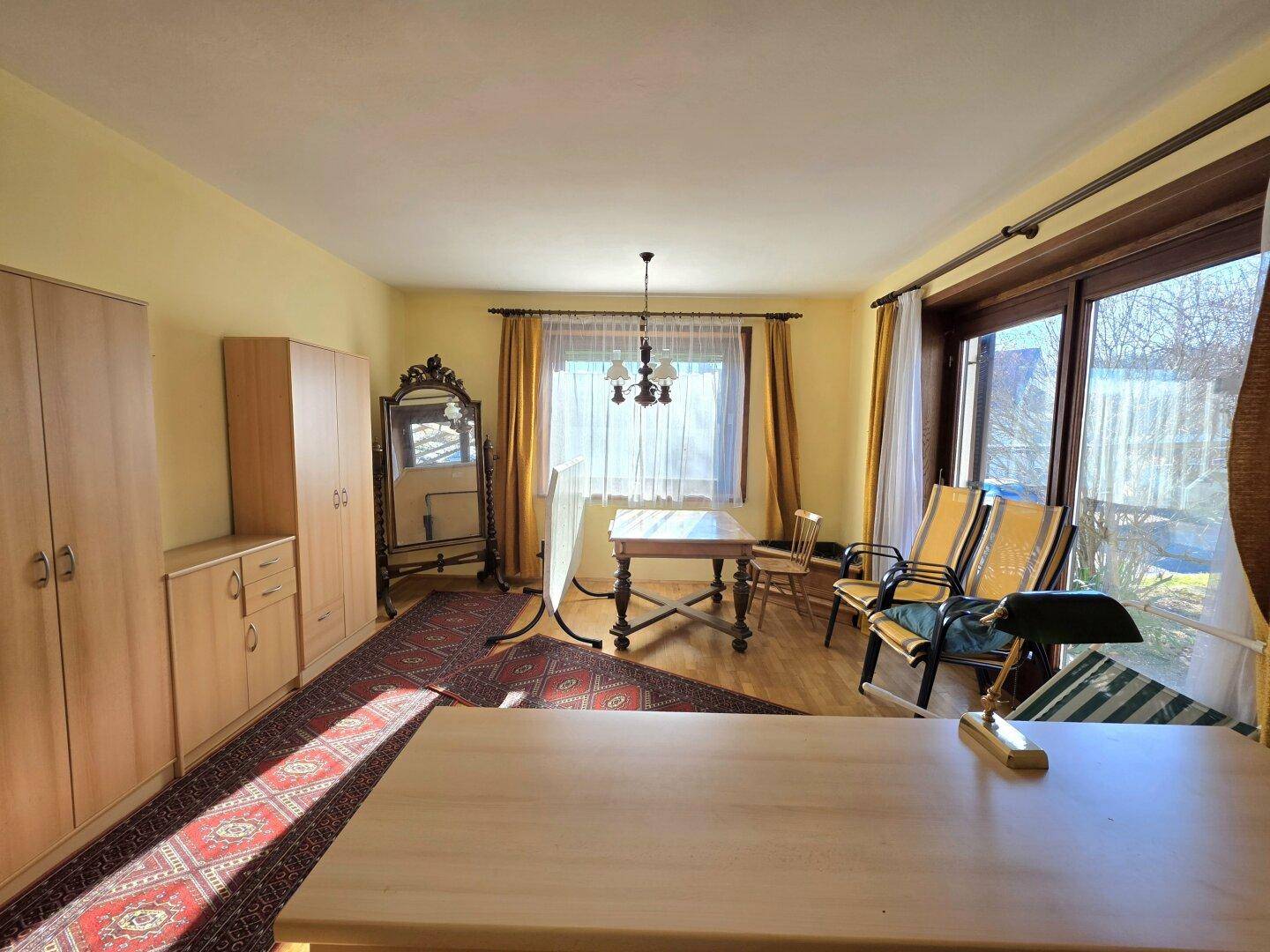 Geräumiger Wohnbereich mit Holzmöbeln, Teppich und großen Fenstern für viel Tageslicht.