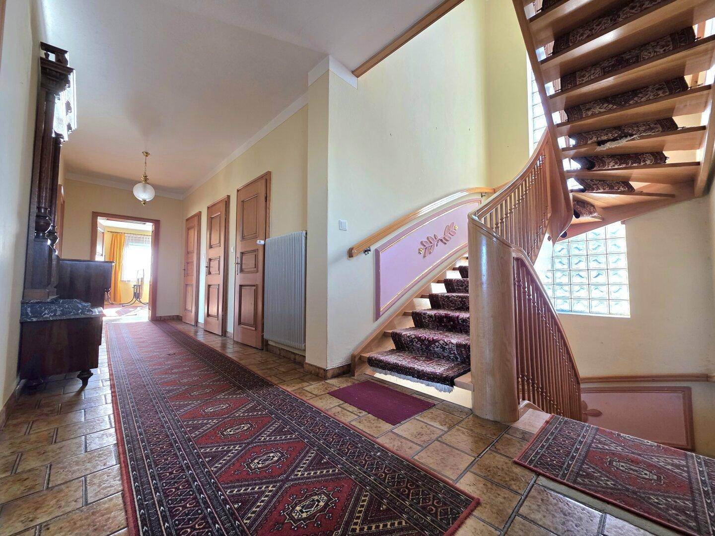 Heller Flur mit geschwungener Holztreppe, gemustertem Teppich und Fliesenboden.
