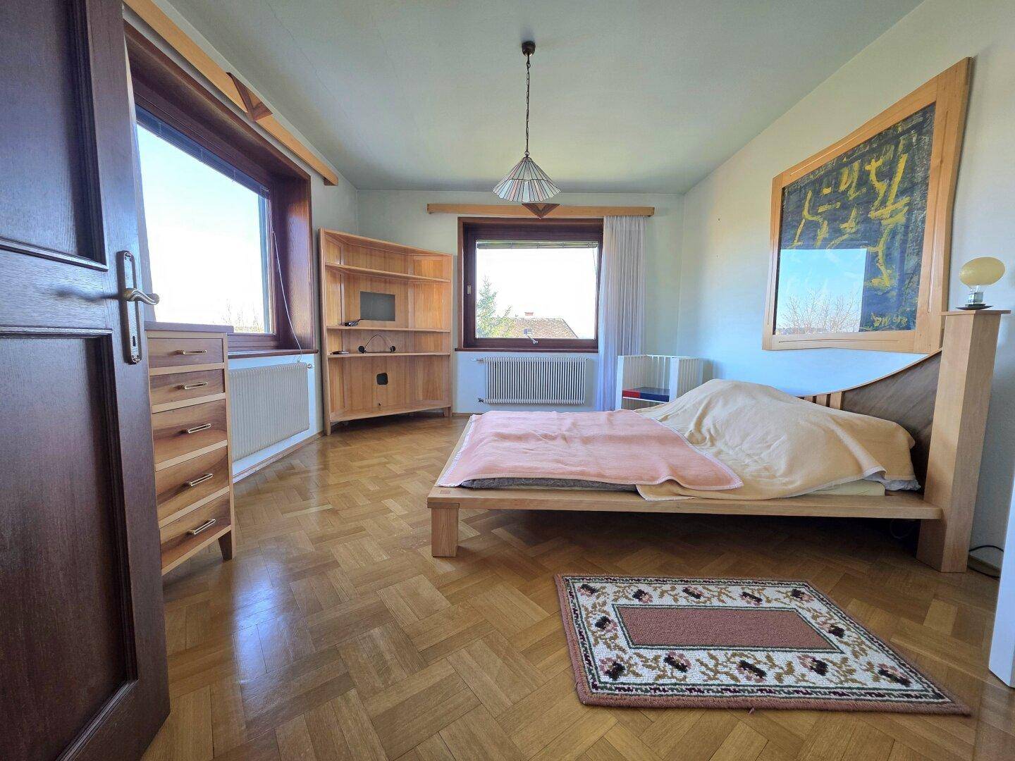 Helles Schlafzimmer mit Holzbett, Parkettboden und großen Fenstern mit Ausblick.