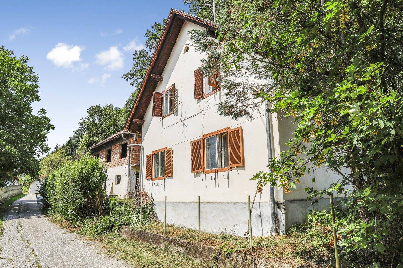 Älteres Haus mit weißer Fassade und braunen Fensterläden, umgeben von viel Grün und einem Feldweg.