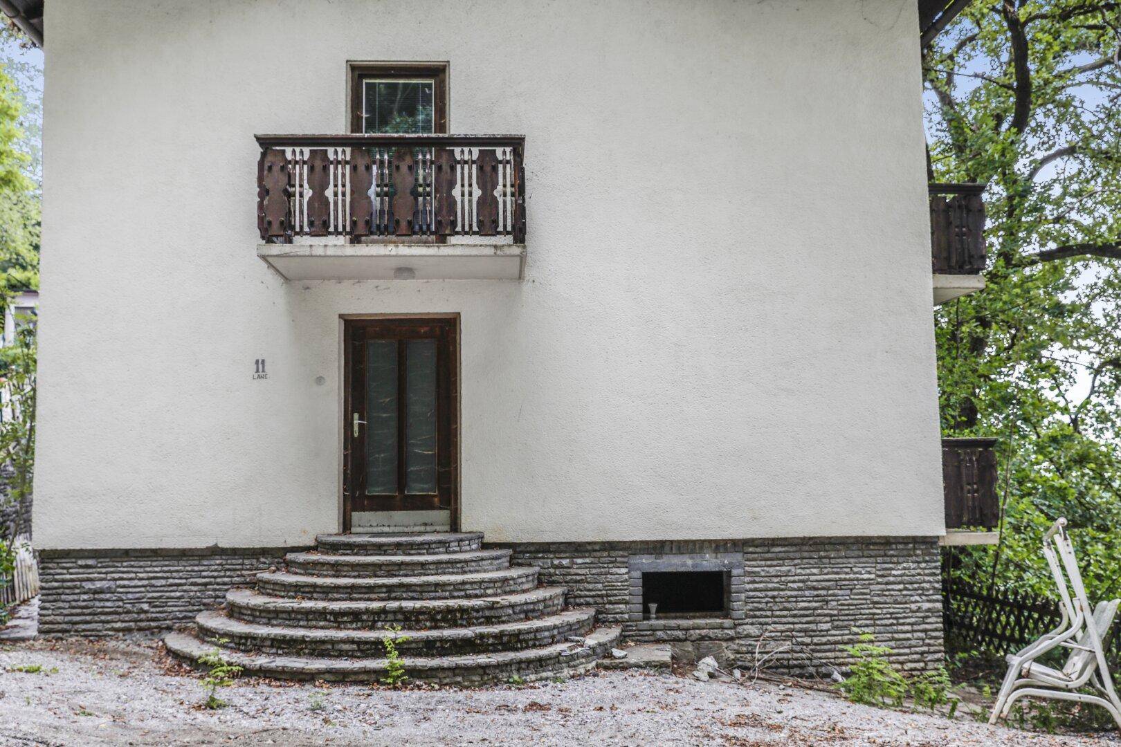 Haupteingang des Hauses mit geschwungenen Steinstufen, Holztür und kleinem Balkon darüber.