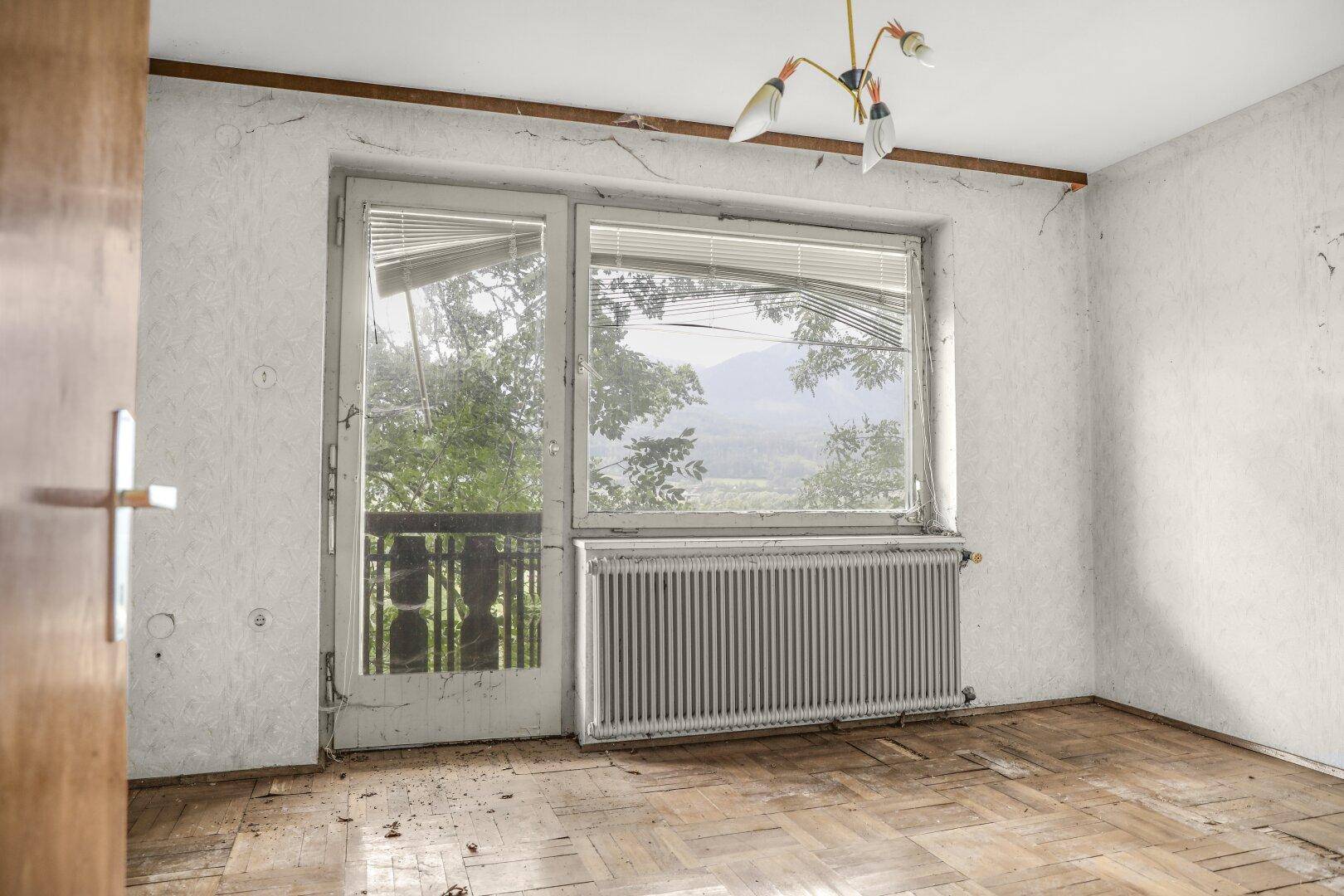 Leeres Zimmer mit großem Fenster, Zugang zu einem kleinen Balkon und einem Heizkörper.