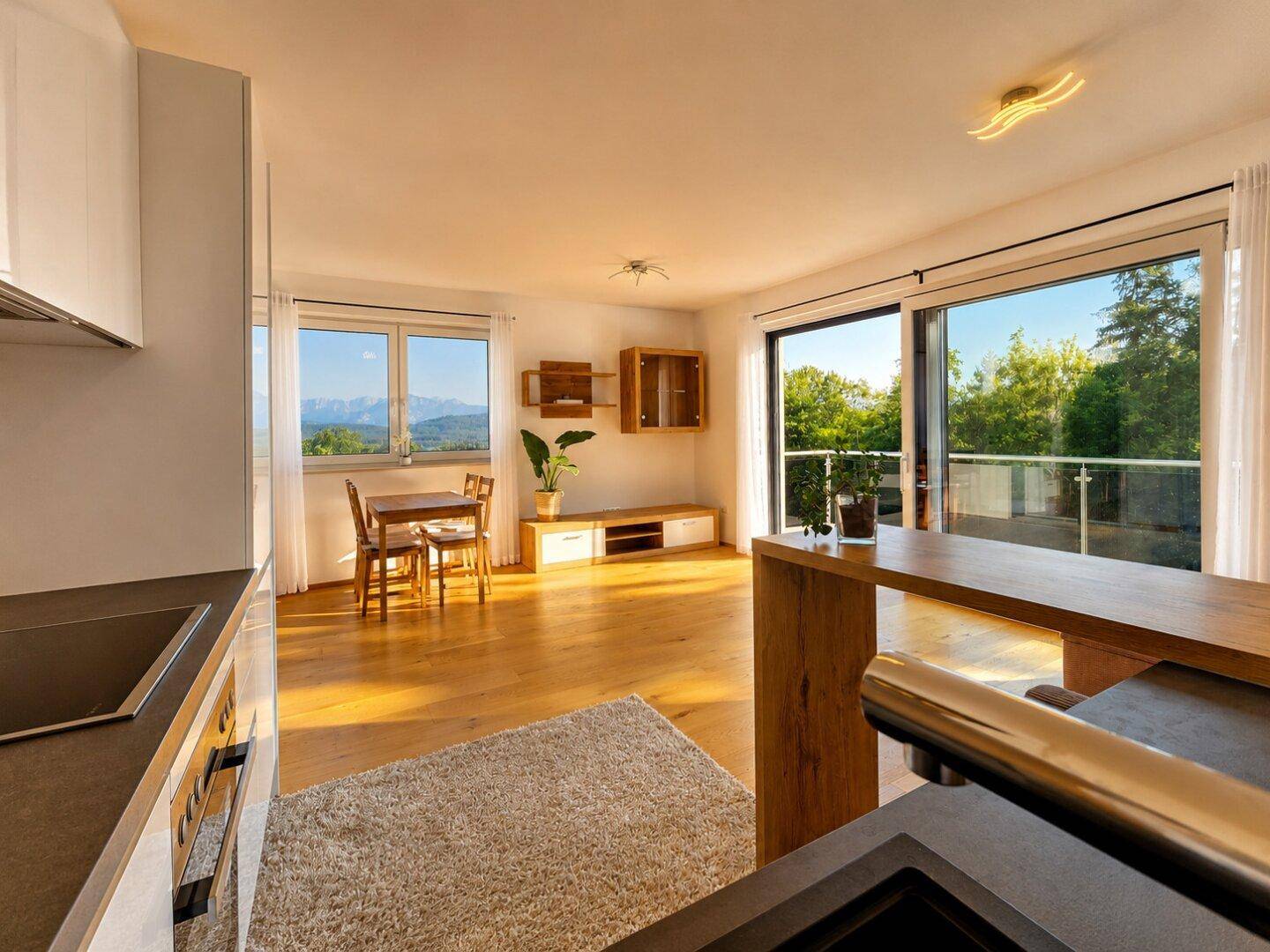 Heller offener Wohn-Essbereich mit Holzboden, integrierter Küchenzeile und Zugang zum Balkon mit Bergblick.