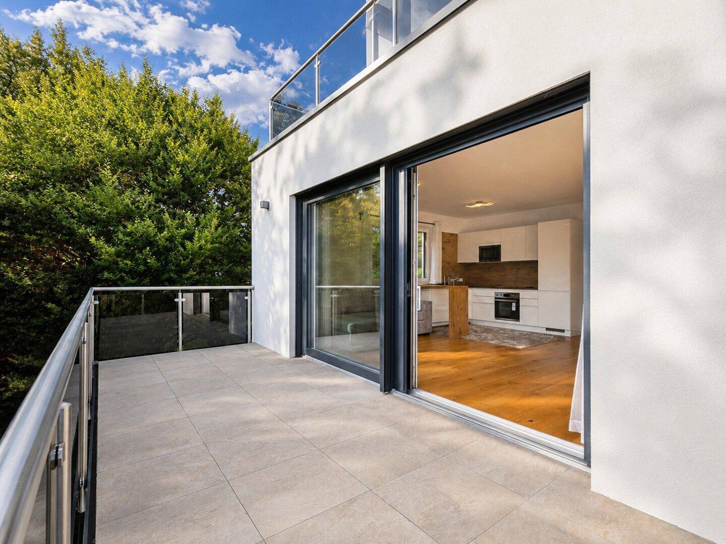 Moderner Balkon mit Fliesenboden und Glasgeländer, bietet Zugang zu einem hellen Wohnbereich mit Küche.