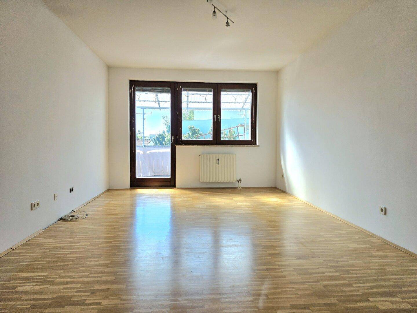 Helles Wohnzimmer mit Parkettboden und großem Fenster, das Zugang zu einem Außenbereich bietet.