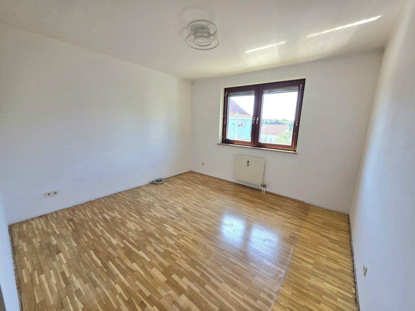 Helles Schlafzimmer mit Parkettboden und Fenster, das viel Tageslicht hereinlässt.