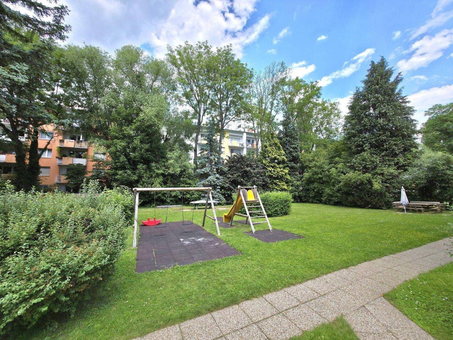 Kinderspielplatz im Gemeinschaftsgarten mit Schaukeln und Rutsche, umgeben von viel Grün.