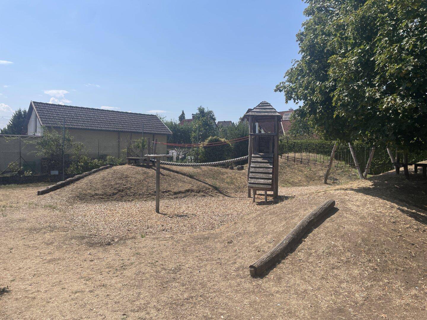 Spielplatz mit Holzturm, Seilbrücke und Schaukeln in einer natürlichen Umgebung.