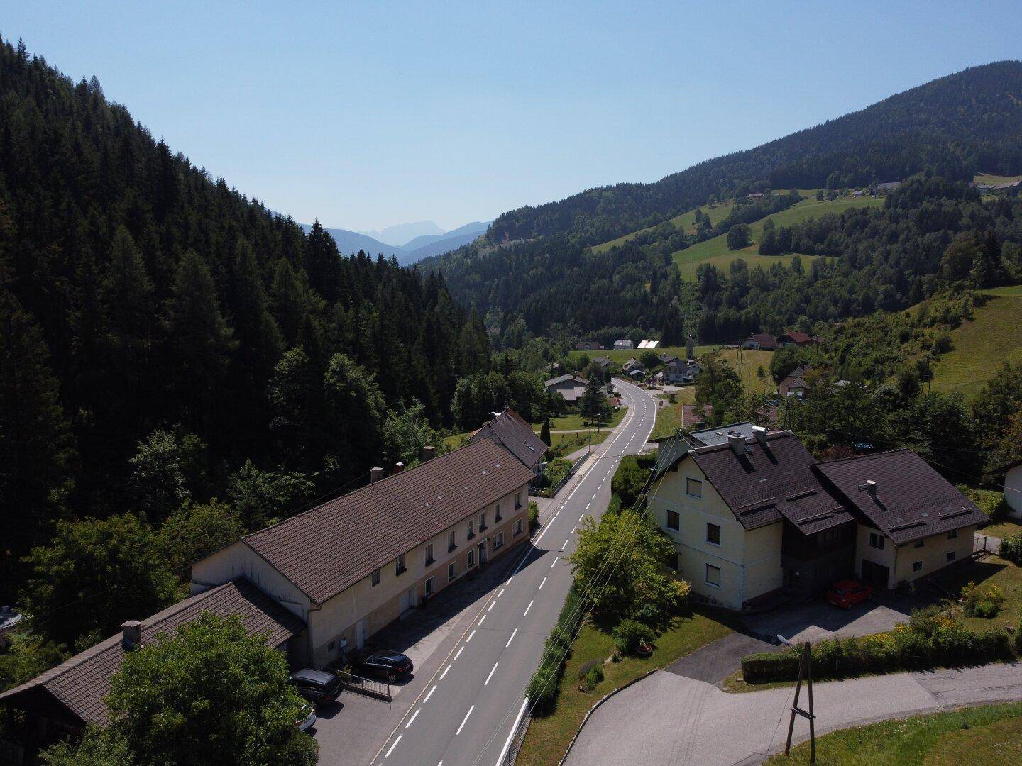Weitläufige Luftaufnahme eines Tals mit einer Straße, umgeben von bewaldeten Bergen und ländlicher Bebauung.