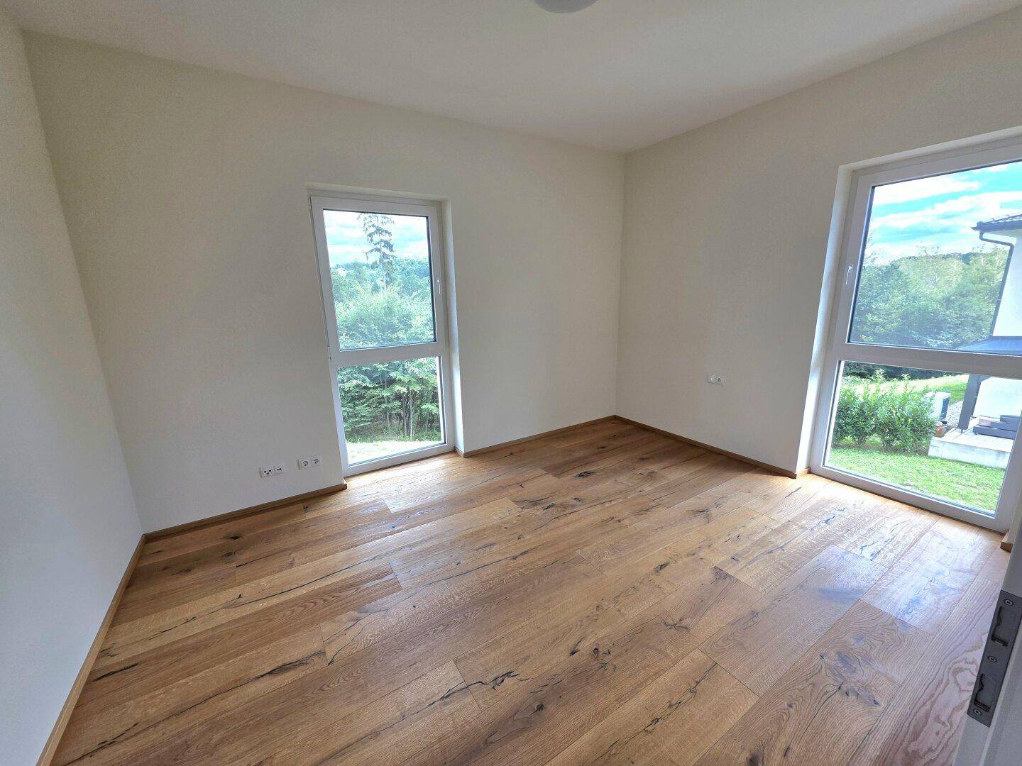 Geräumiges Schlafzimmer mit Holzboden und zwei Fenstern mit Blick ins Grüne.