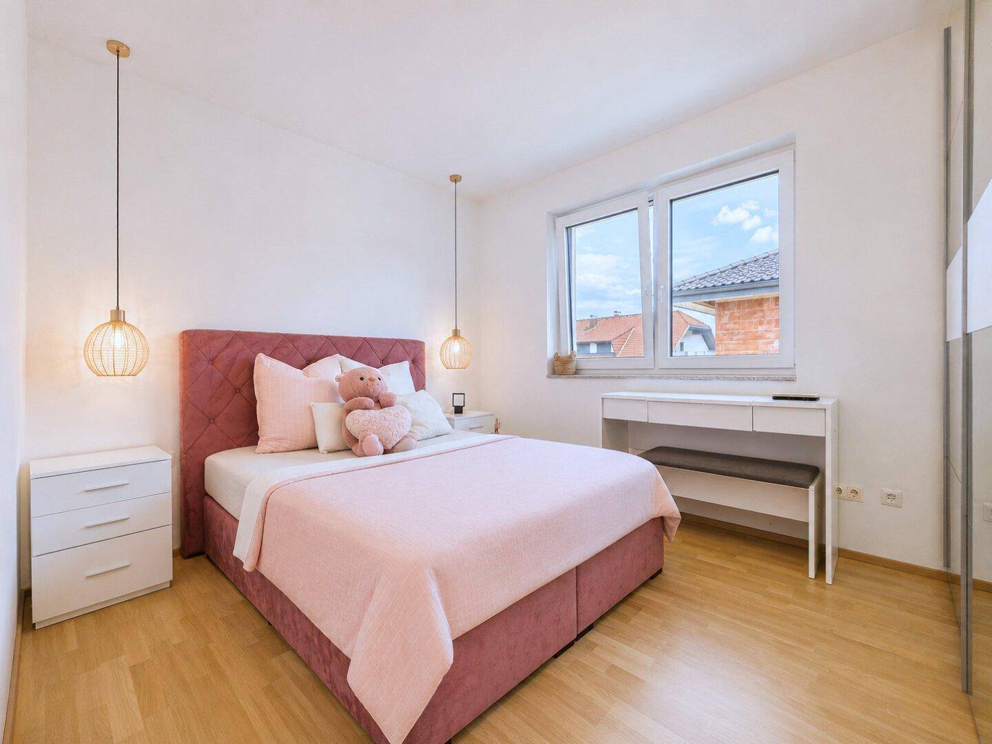 Gemütliches Schlafzimmer mit rosa Polsterbett, Nachttischen und Schreibtisch am Fenster.
