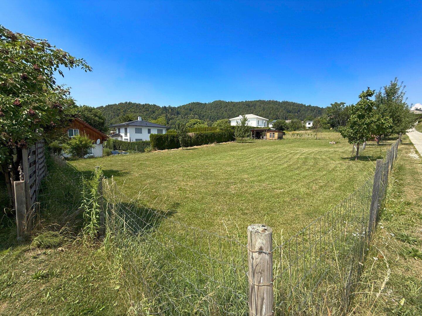 Großes, eingezäuntes Gartengrundstück mit Rasenfläche und Wohnhäusern sowie Hügeln im Hintergrund.