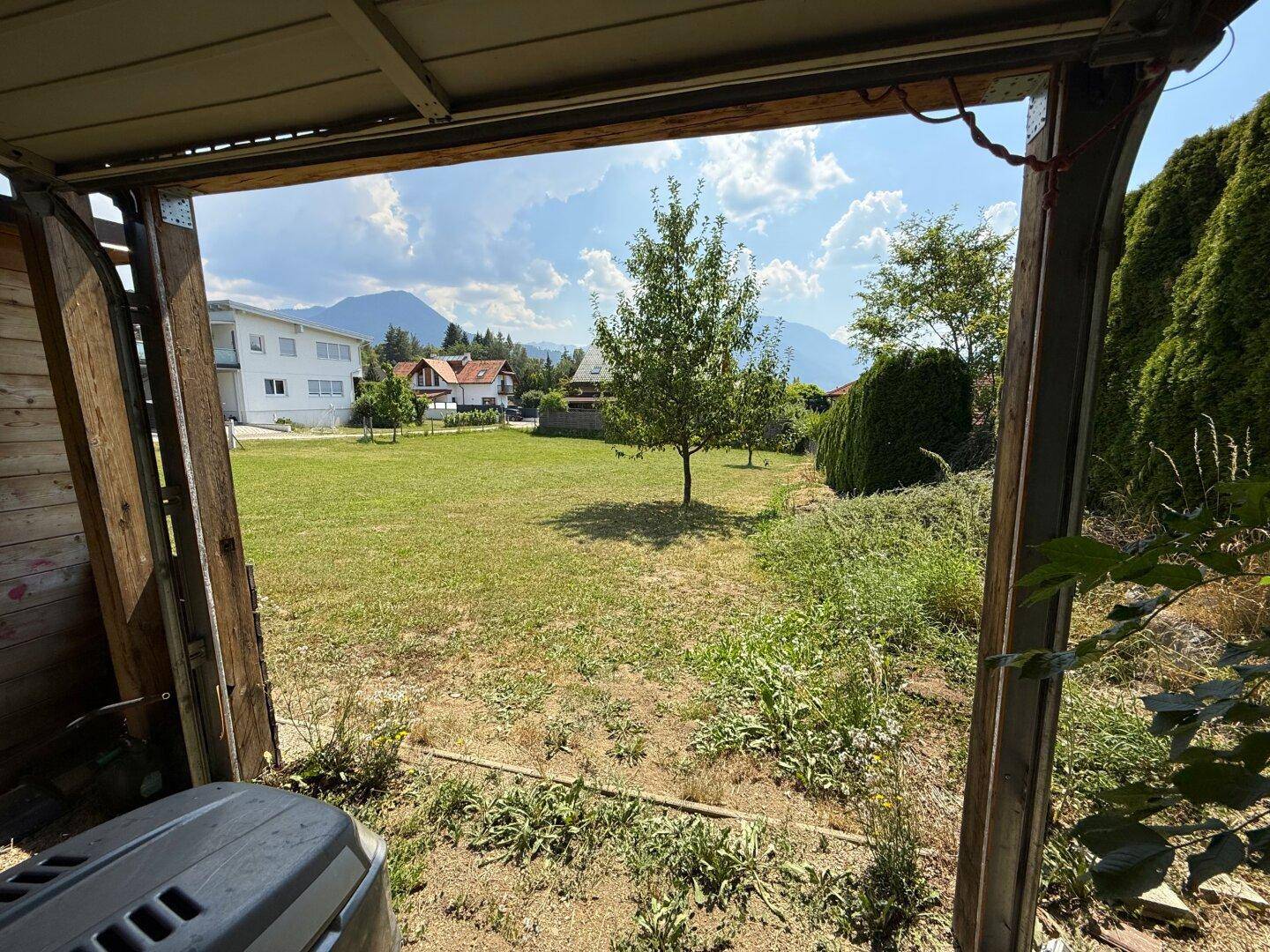 Blick aus einem Holzschuppen auf eine grüne Rasenfläche mit Häusern und Bergen in der Ferne.