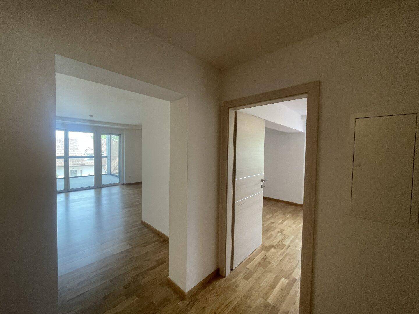 Flur mit Holzboden und Blick durch eine offene Tür in einen hellen Wohnbereich mit Balkonzugang.