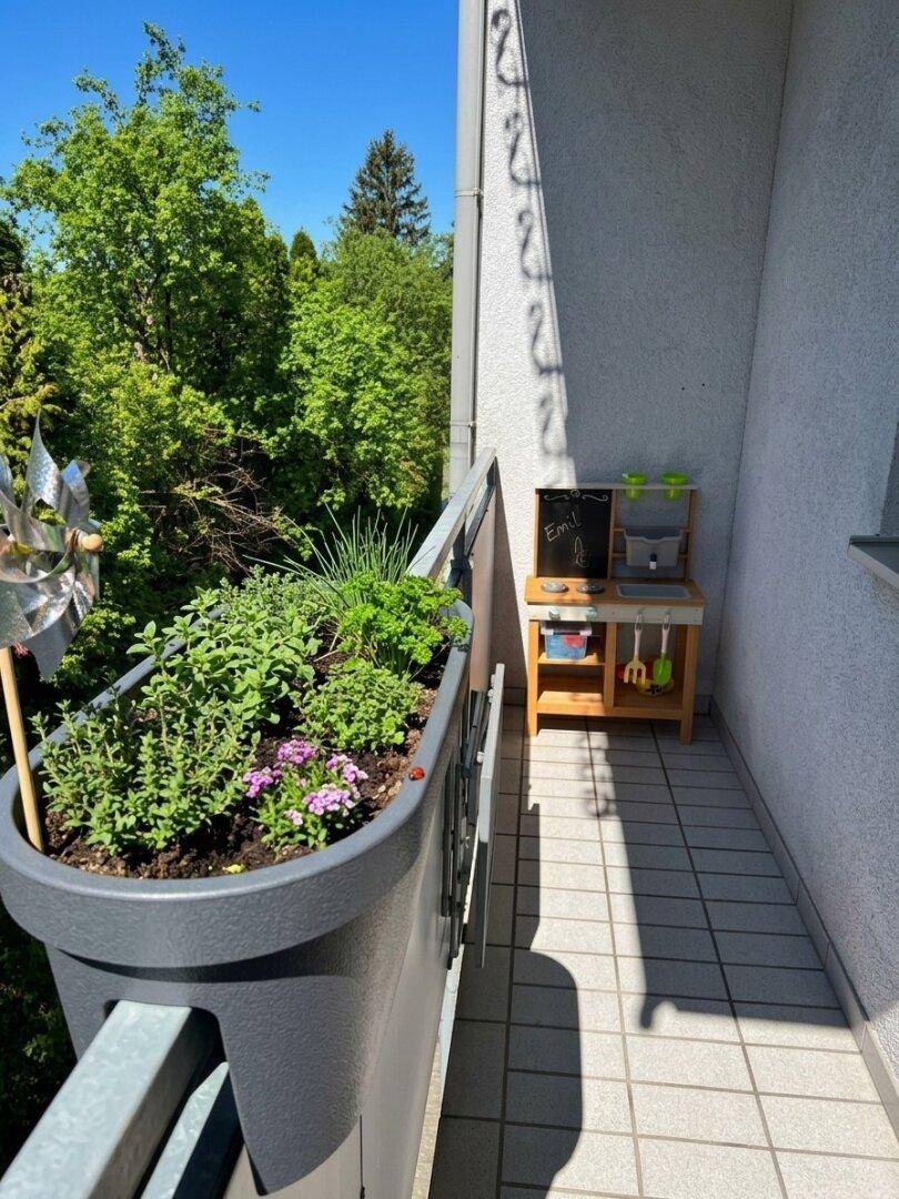 Schmaler Balkon mit Pflanzenkübeln und einer kleinen Spielküche für Kinder im Freien.