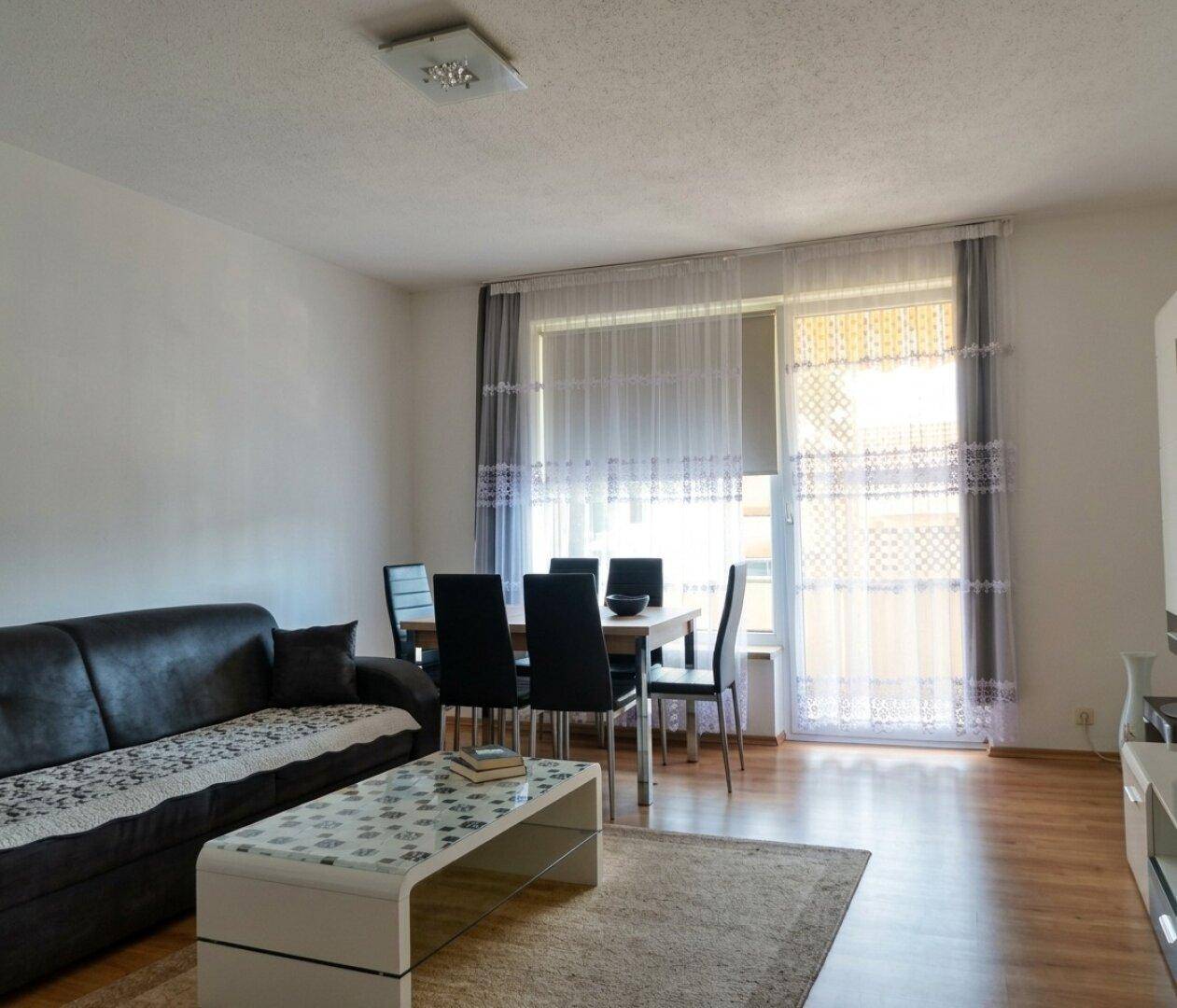 Gemütlicher Wohn-Essbereich mit dunklem Sofa, Esstisch und Zugang zu einem hellen Außenbereich.
