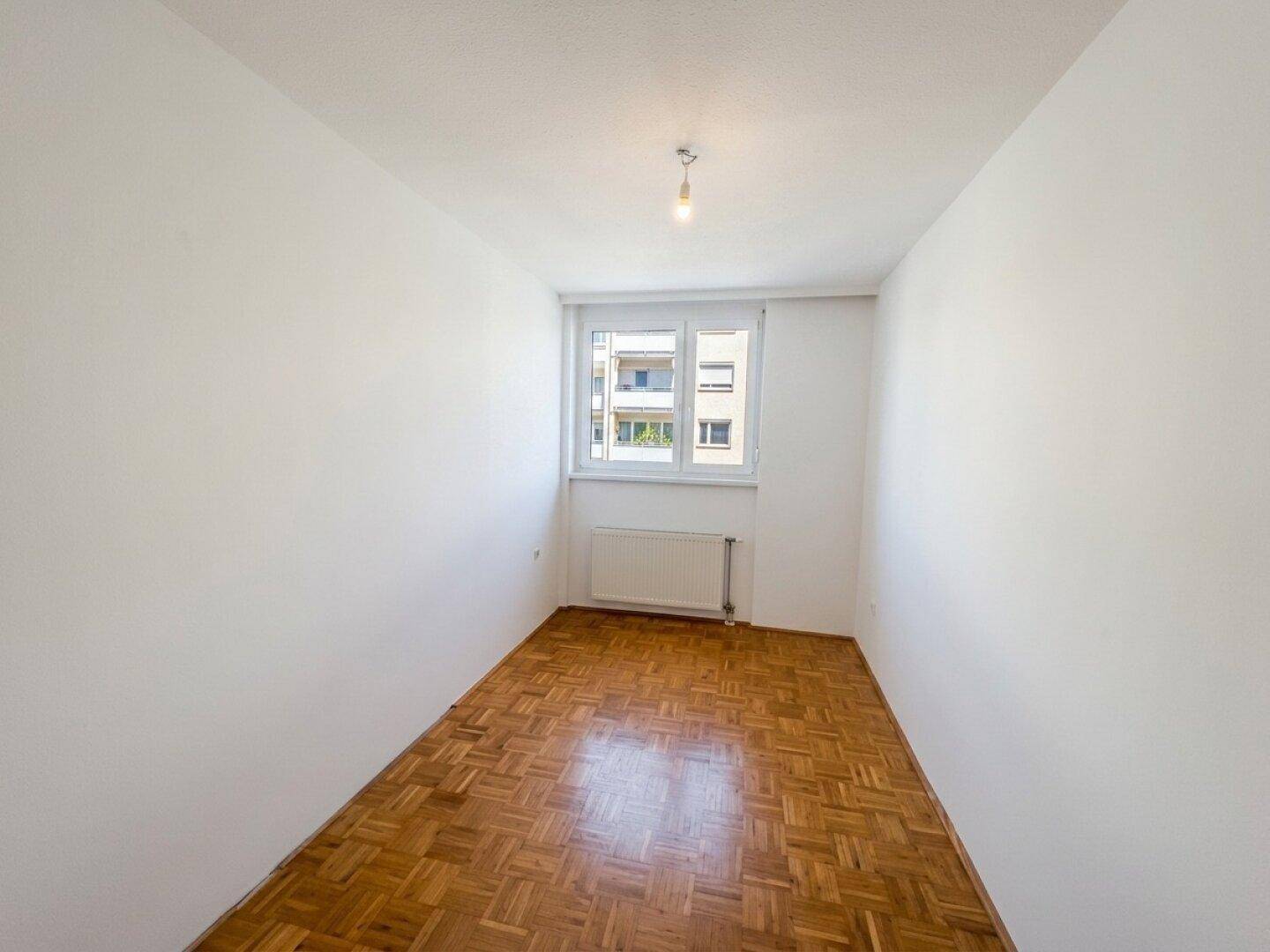 Leeres Zimmer mit Parkettboden und Fenster, ideal als Schlaf- oder Arbeitszimmer.