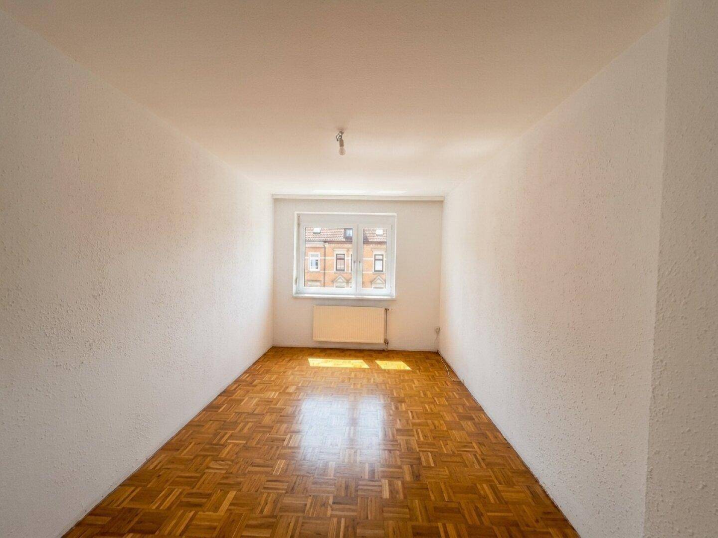 Helles, leeres Zimmer mit Parkettboden und Fenster.