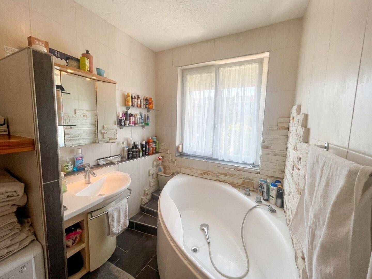 Geräumiges Badezimmer mit großer ovaler Badewanne, Waschbecken und Fenster, hell gefliest.