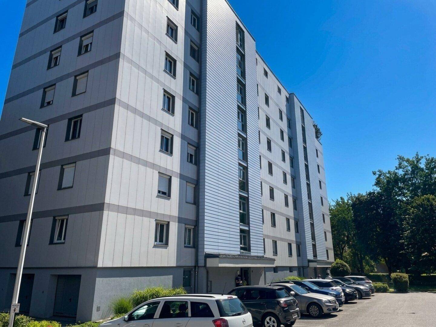 Mehrstöckiges Wohngebäude mit moderner Fassade und zahlreichen Parkplätzen im Freien.
