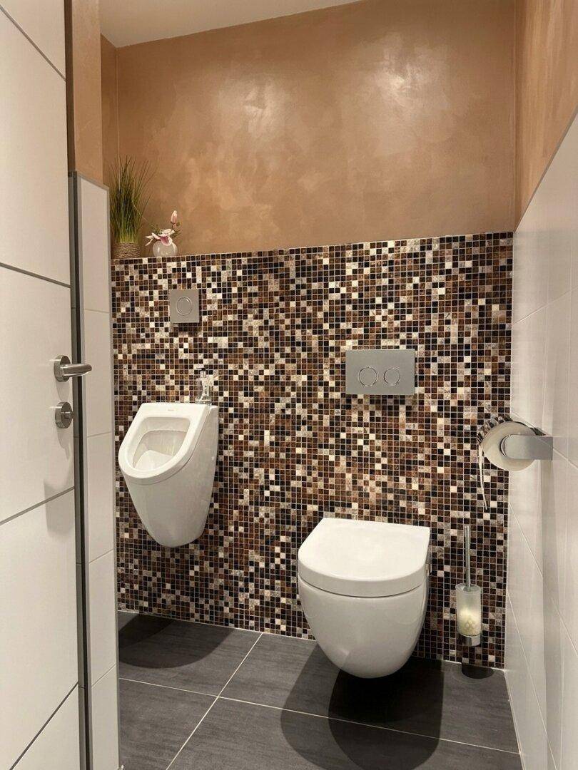 Modernes WC mit Toilette, Urinal und dekorativen Mosaikfliesen an der Wand.